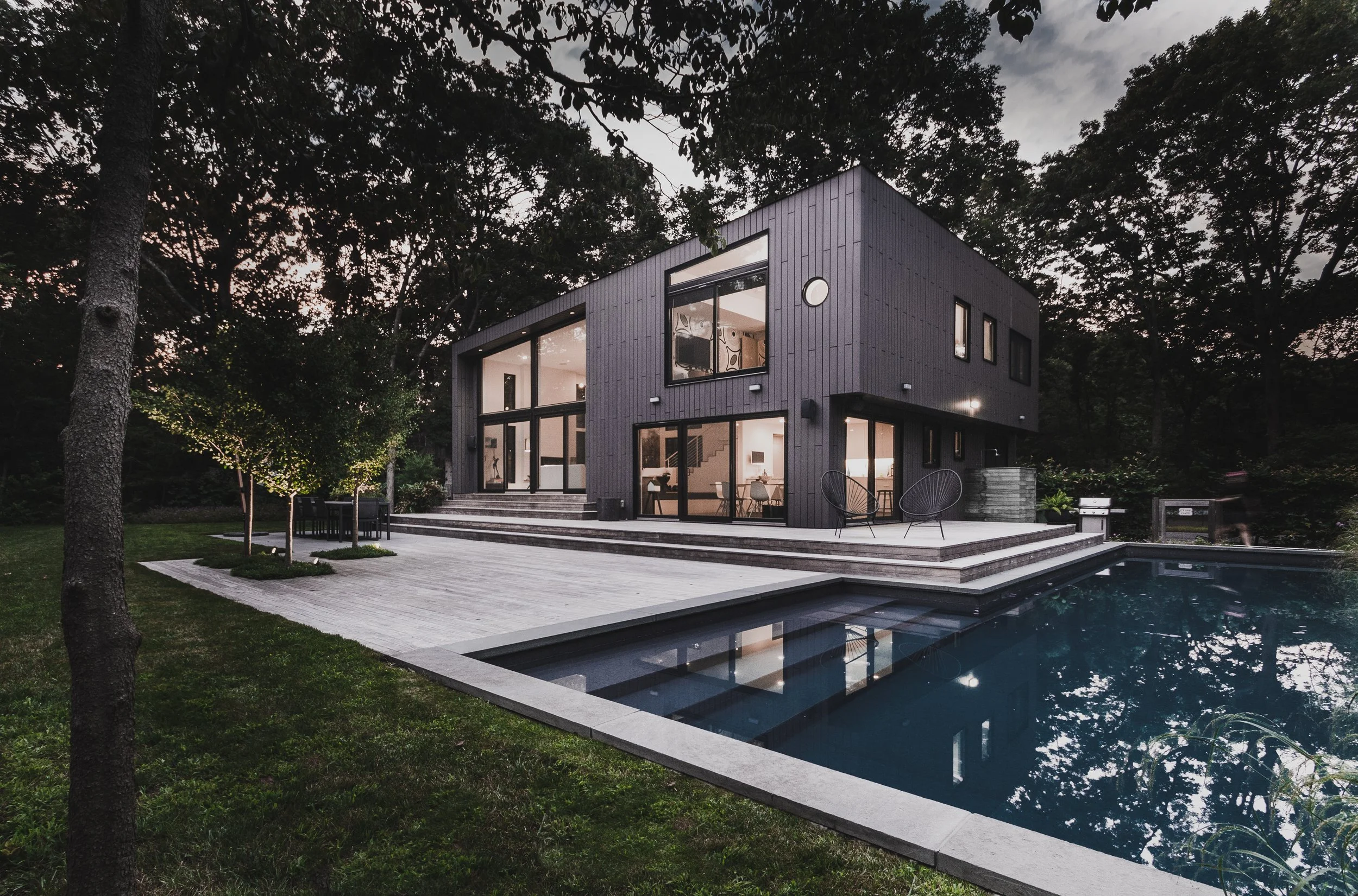 Modern black house with large windows, deck, and swimming pool, surrounded by trees.