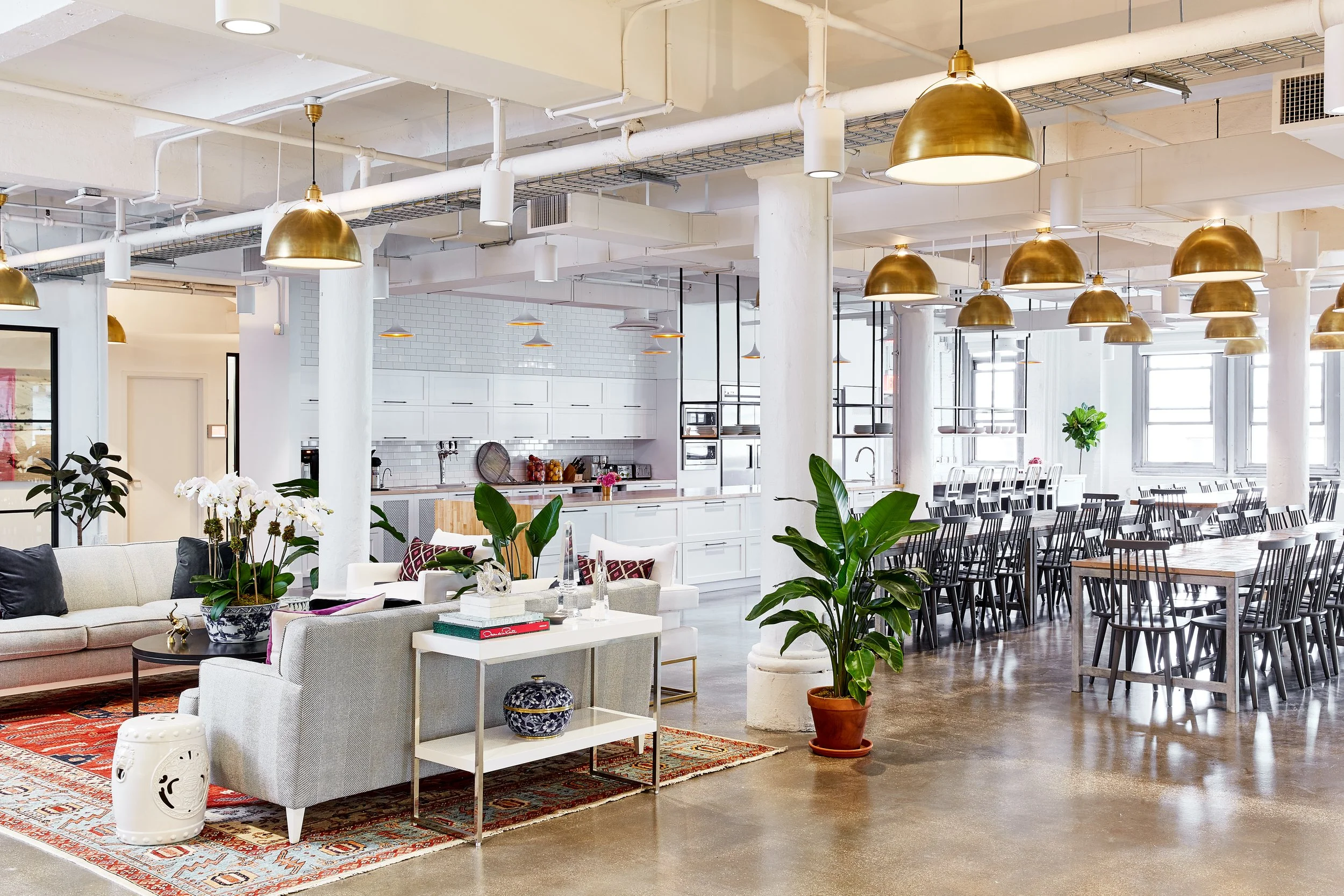 Modern open-plan office space with a kitchen, dining tables, and seating area. Features white cabinetry, wooden tables, black chairs, couches, plants, and overhead gold pendant lights.