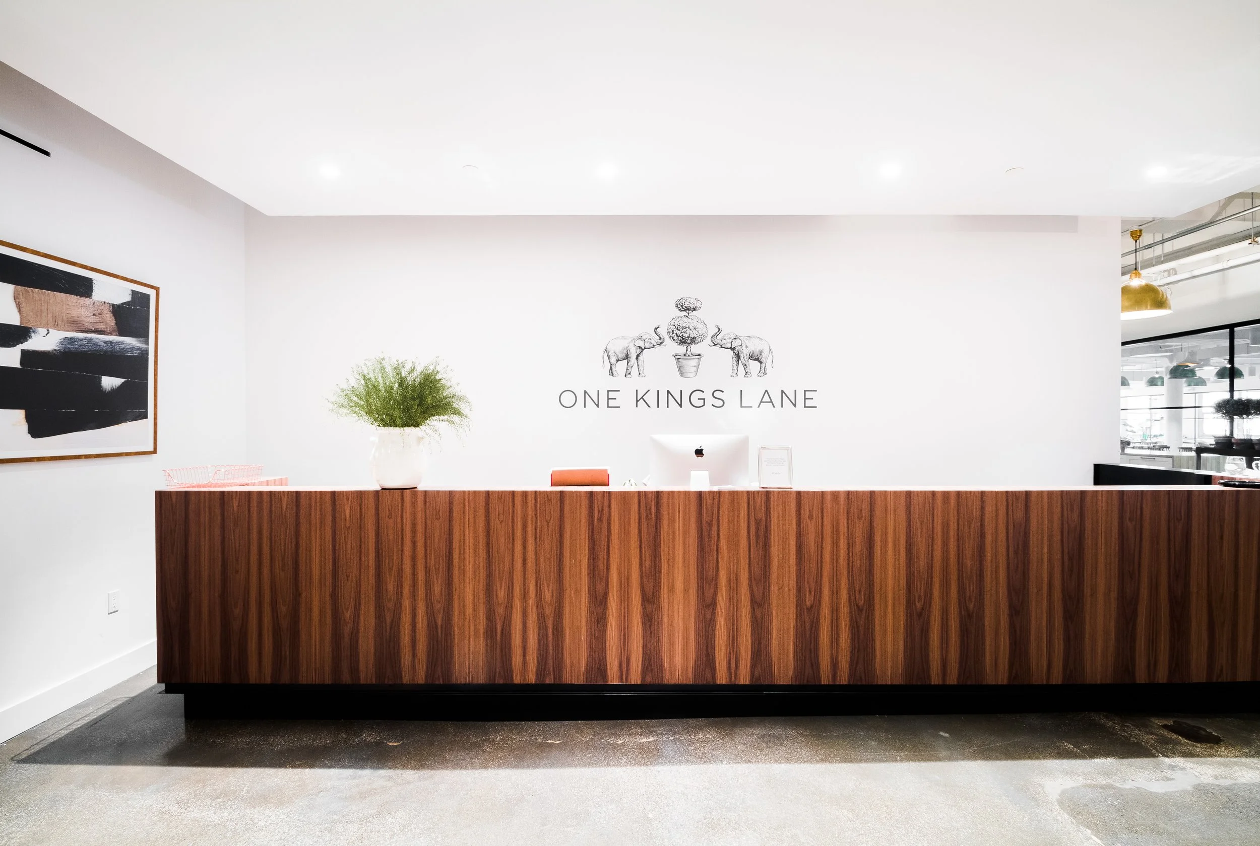 A reception desk at One Kings Lane with a wooden counter, a potted plant, a computer, and a wall logo featuring two elephants and a topiary. Artwork is displayed on the wall to the left.