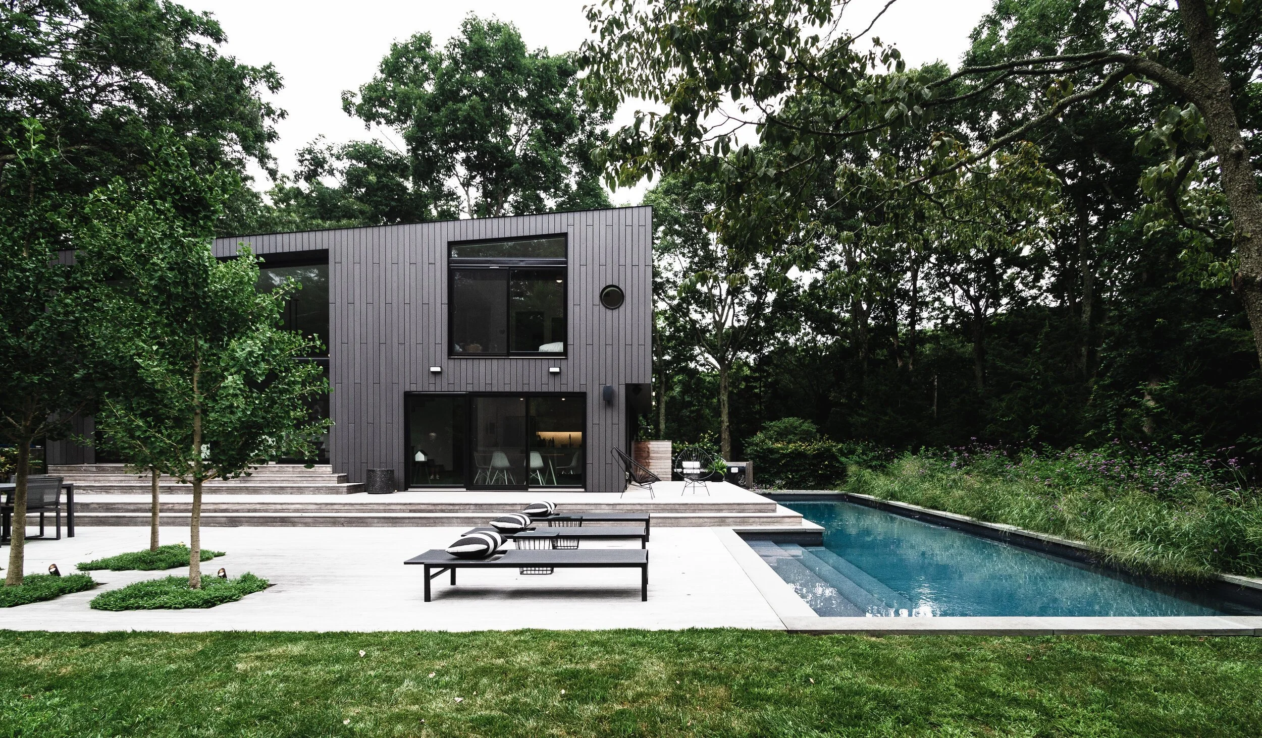Modern two-story house with a black exterior, patio, swimming pool, and surrounding trees.