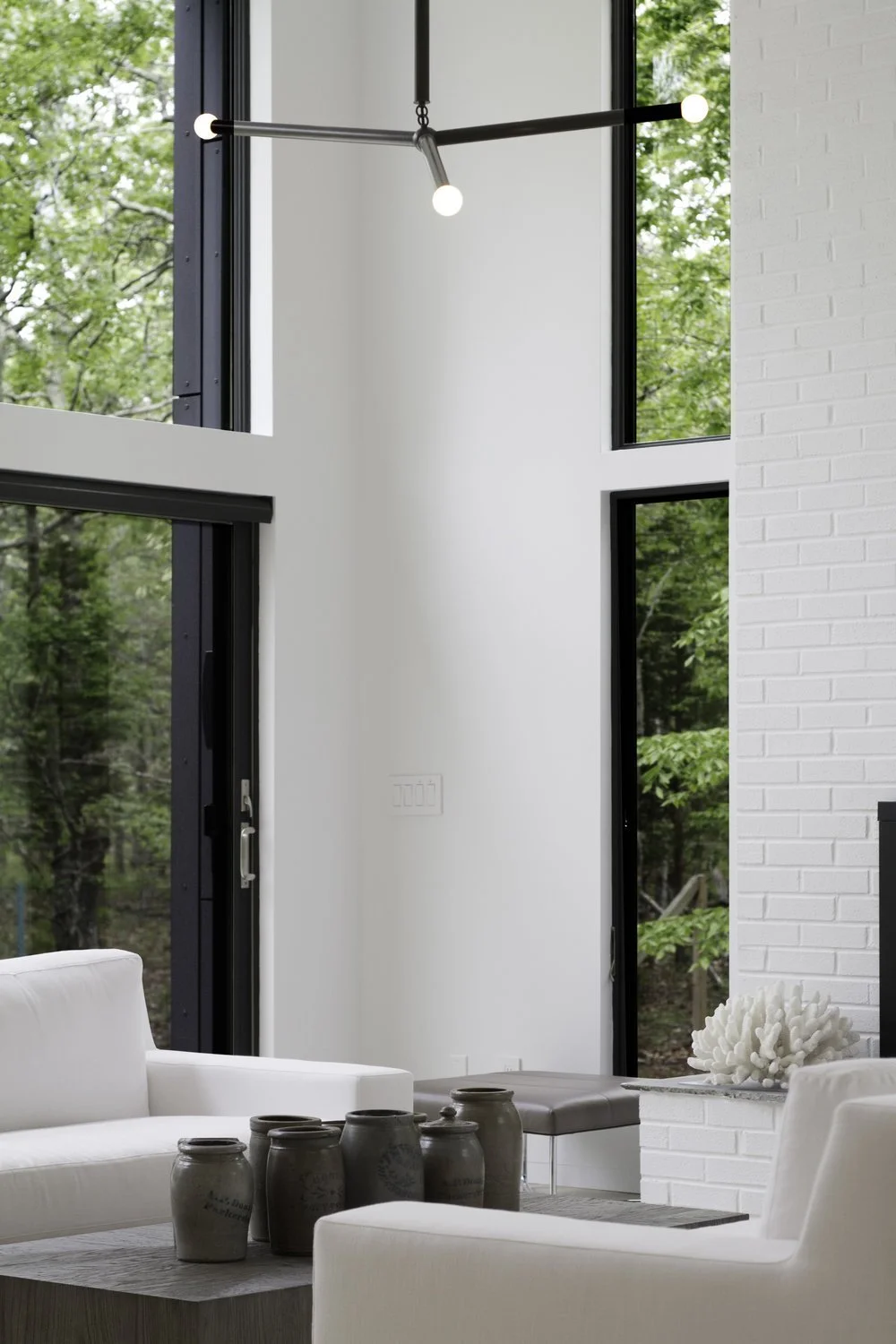 Modern living room with large windows, white sofa, black chandelier, and decorative jars on a table. The room features a white brick wall and view of green trees outside.