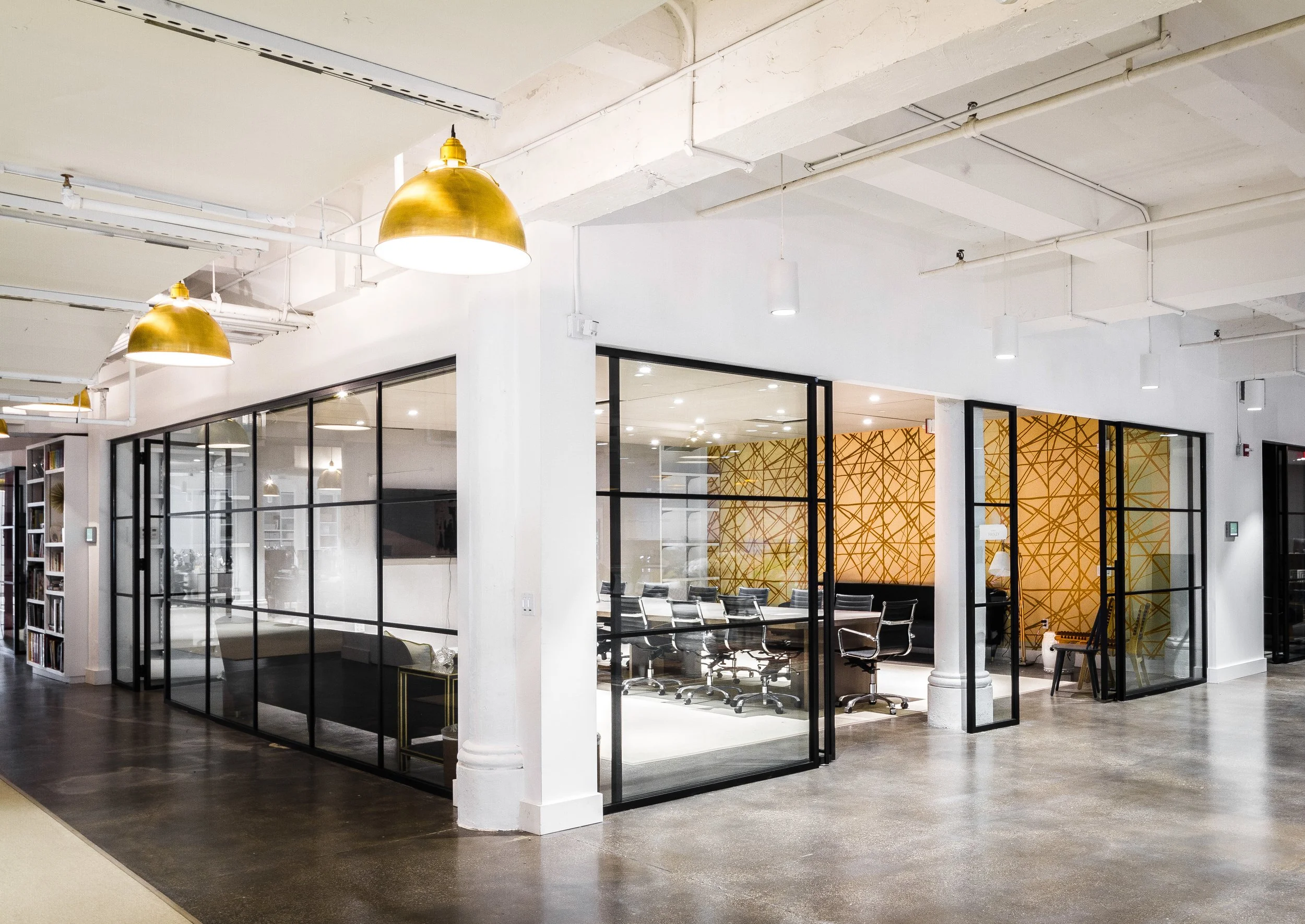 Modern office interior with glass-walled conference room, black chairs, decorative golden wall design, polished concrete floor, and exposed ceiling with gold dome lights.