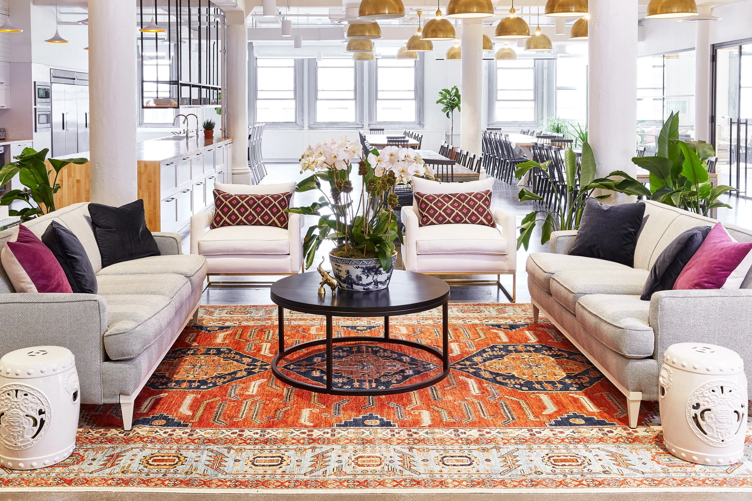 Modern office lounge with sofas, armchairs, decorative pillows, a central black round table with a floral centerpiece, a vibrant red and blue patterned rug, potted plants, and adjacent to an open kitchen area with dining tables and pendant lights.