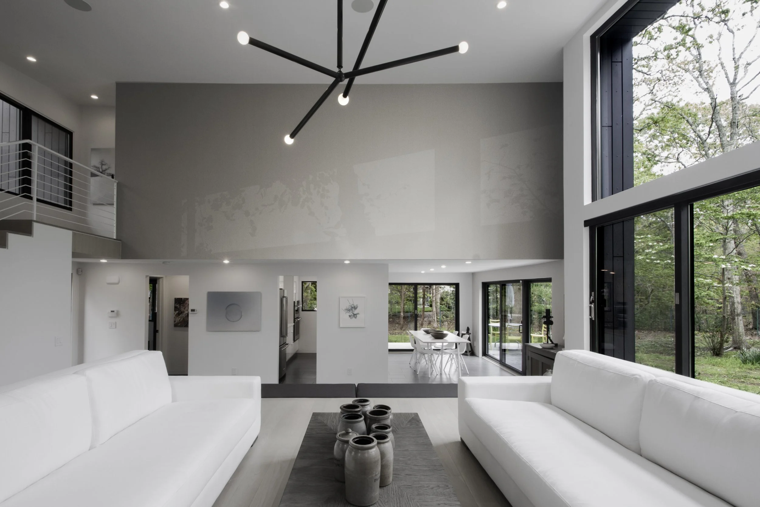 Modern living room with white sofas, contemporary light fixture, large windows, and a view of a dining area and outdoor greenery.