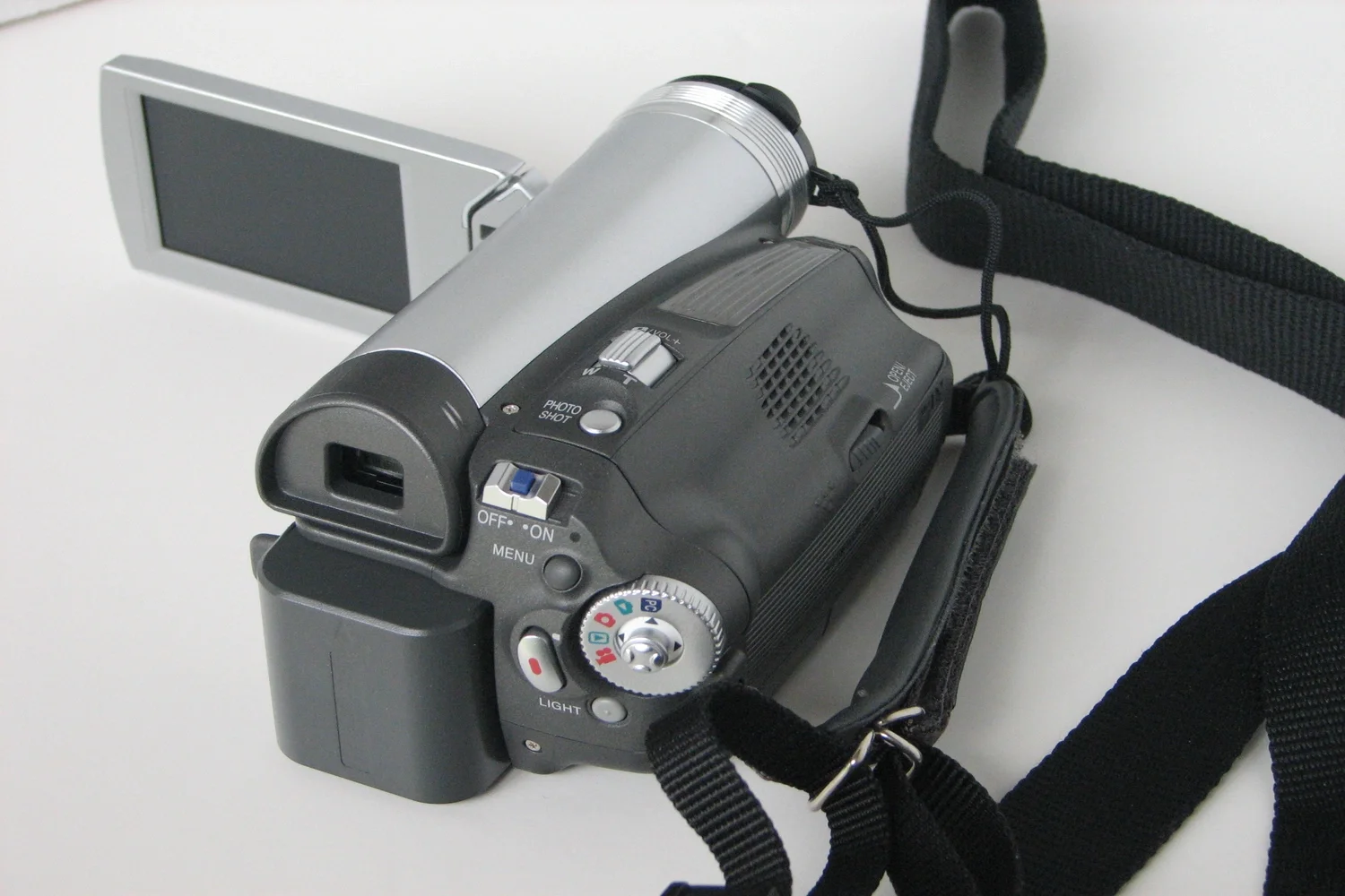 A digital camera sports a black grip and control dial, attached to a black strap, resting on a white surface with a silver flip-out screen extended.