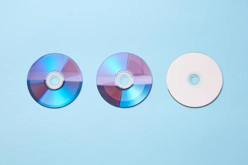Three compact discs, two reflective and one plain, arranged in a row on a light blue background.