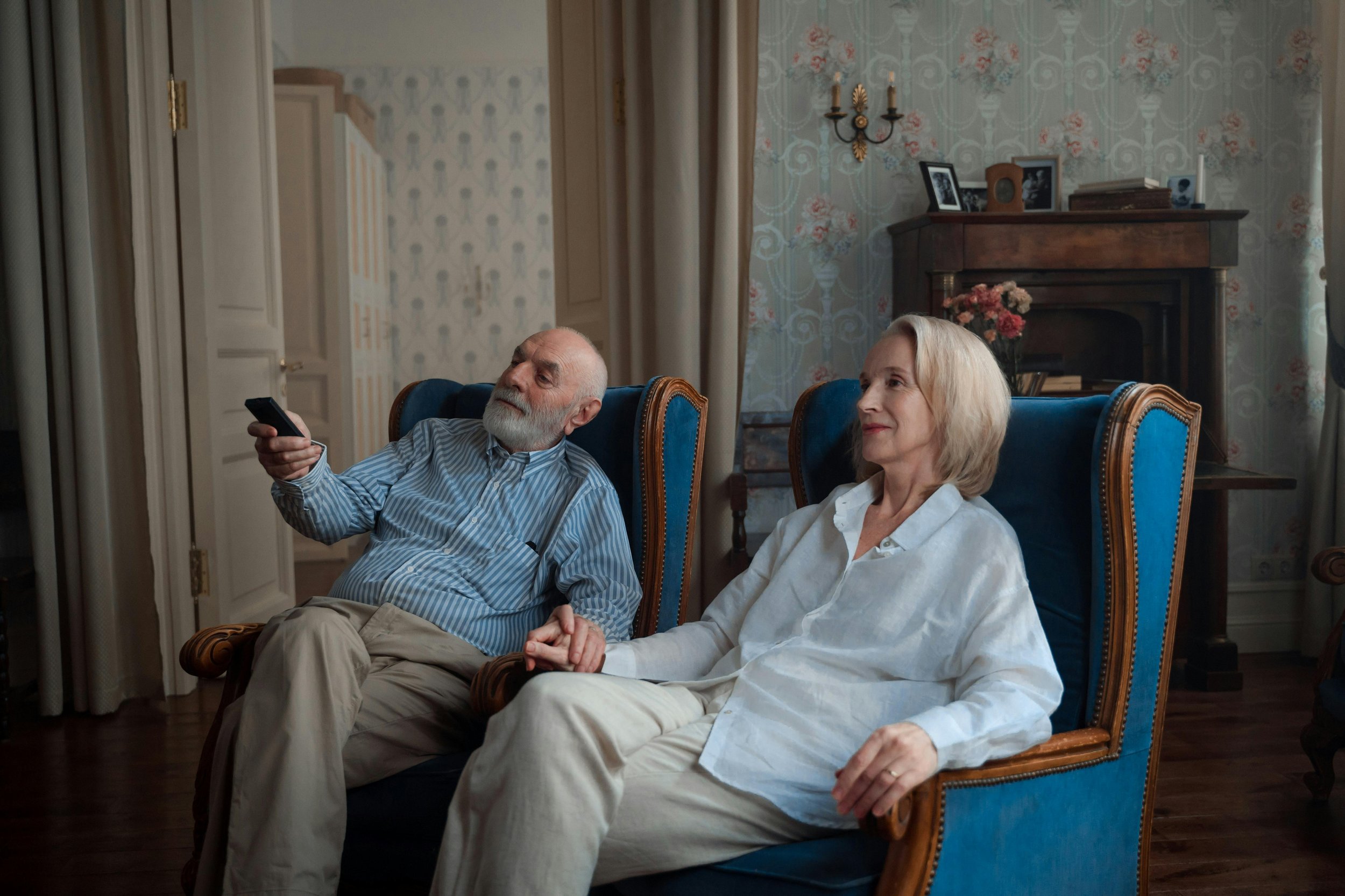 An elderly couple sits together in armchairs, holding hands, with a man holding a remote control. The room has vintage decor, with a fireplace, ornate wallpaper, and framed photos on the mantle.