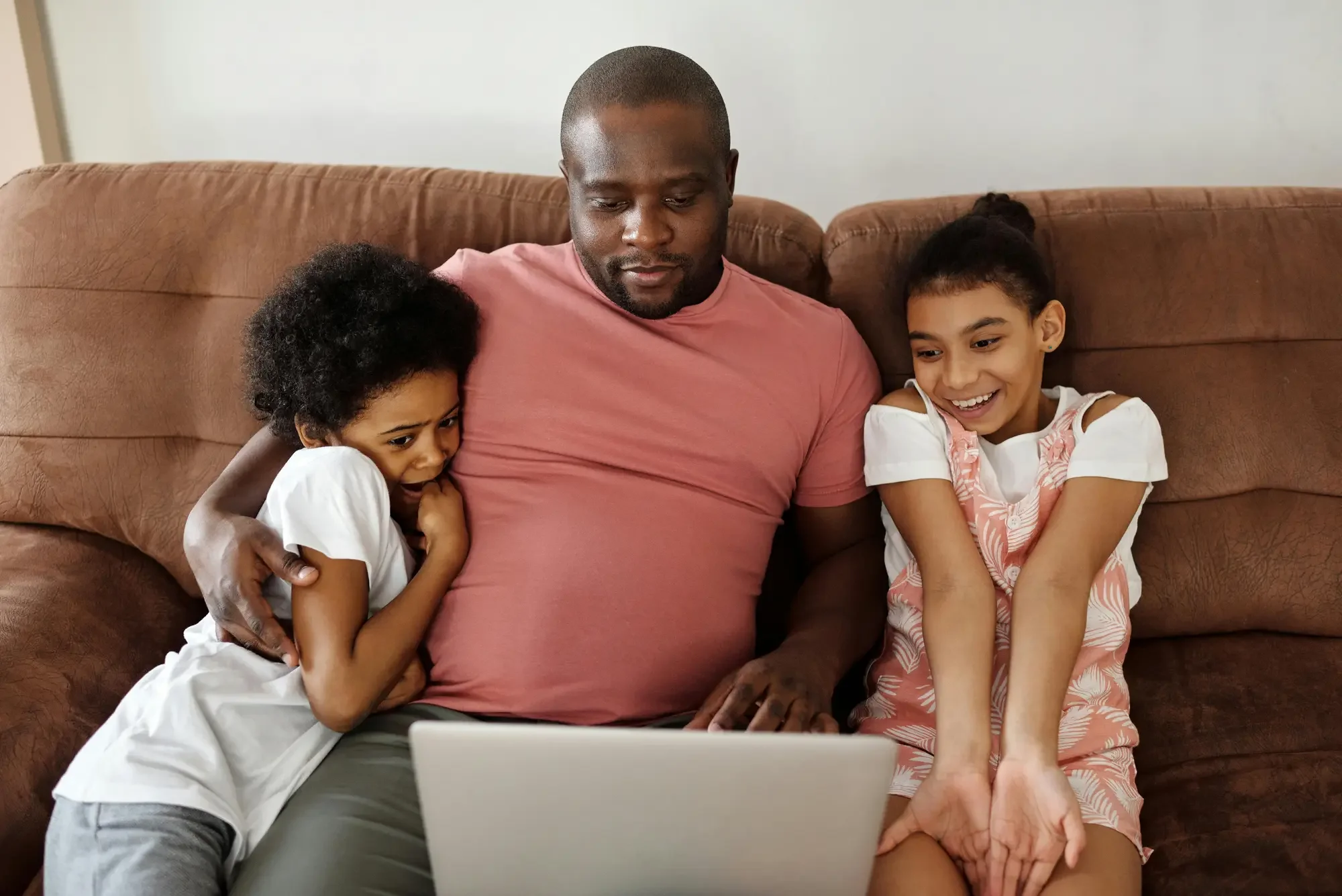 A man with two young girls sitting on a brown couch, watching their digitized videos on a laptop. The girls are excited and smiling.