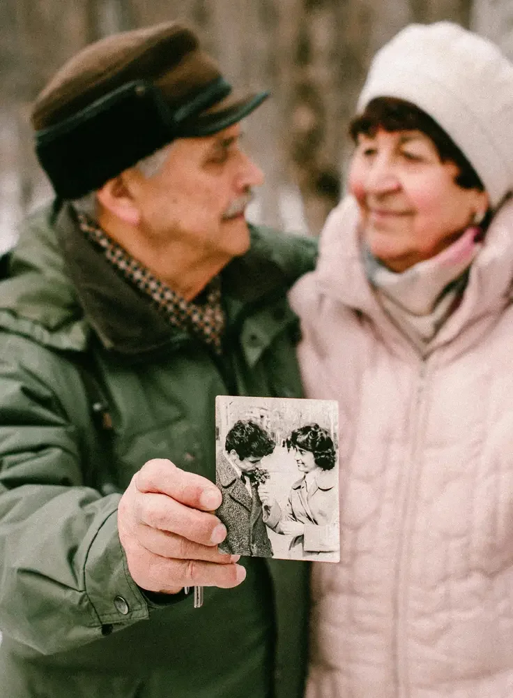 An elderly man and woman are outdoors, with the man holding a black-and-white photograph of a young man and woman. They are smiling at each other