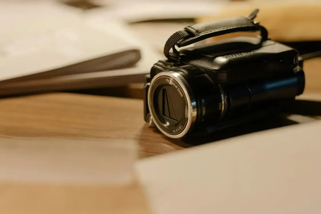 Close-up of a black camera with a lens, placed on a wooden surface near papers and notebooks.