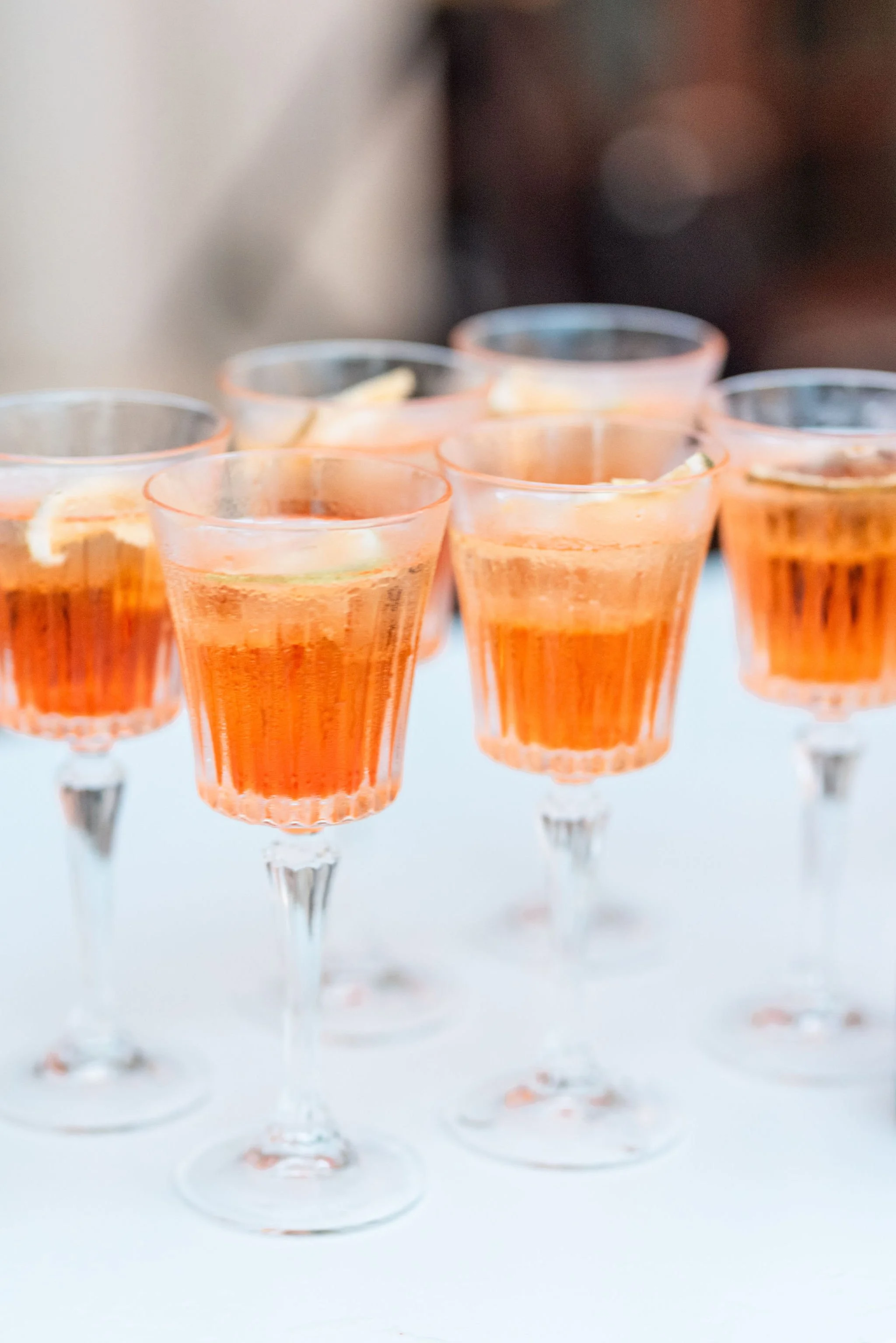Six glasses of orange cocktail with lemon garnishes on a white surface.