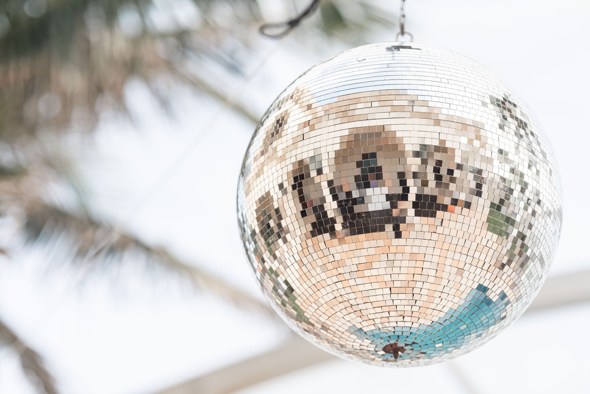 A close-up of a hanging disco ball with a reflective surface, distorting surrounding images and palm trees in the background.