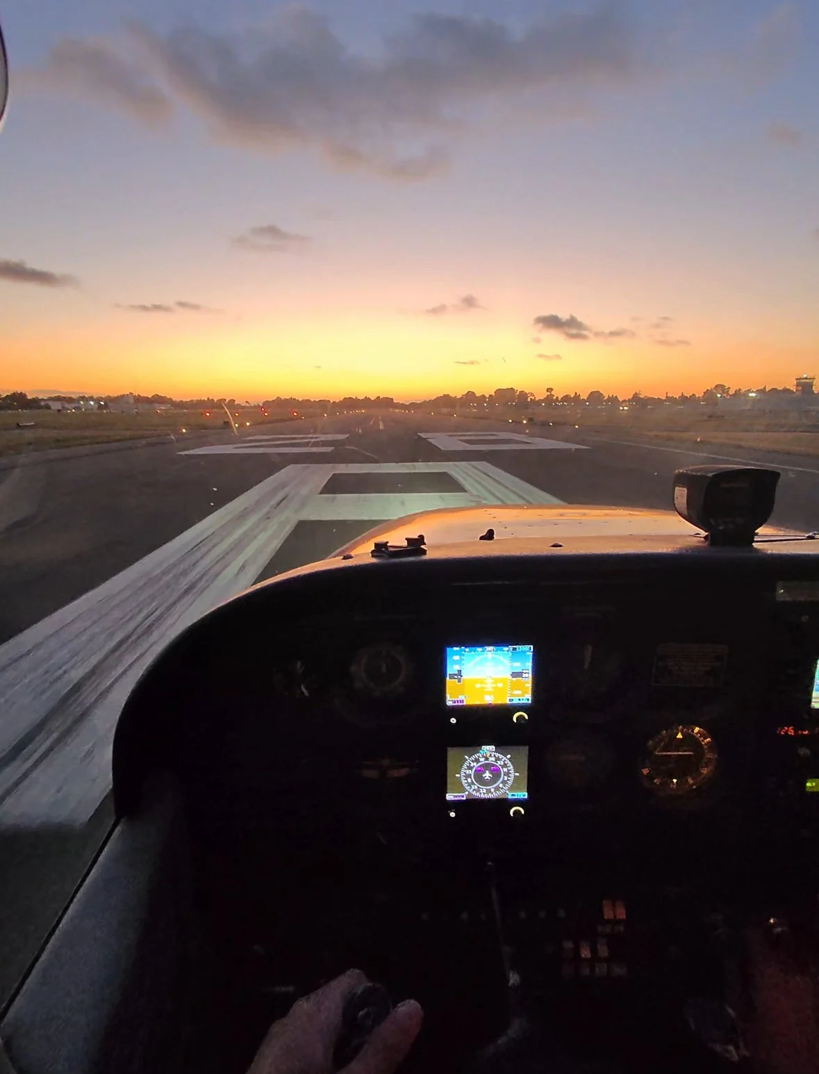 View from inside an airplane cockpit on the ground at sunset, runway ahead, with a sunset sky and scattered clouds.