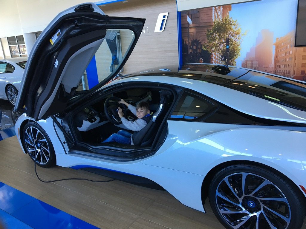 A young boy sitting inside a white BMW i8 sports car with the door open, inside a showroom.