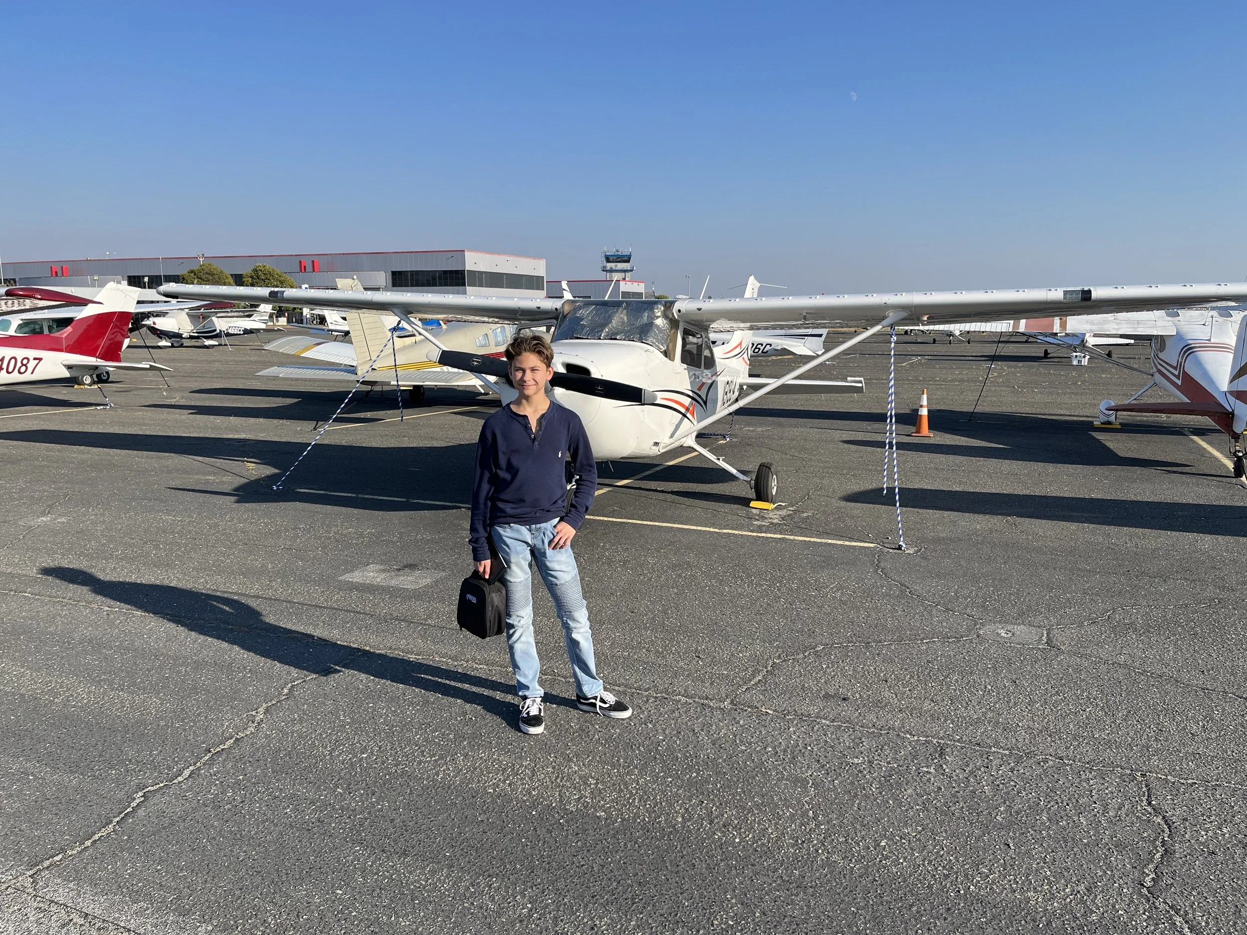 A young boy standing in front of small airplanes on an airport tarmac holding a black bag. The airplanes are parked and secured with ropes, with a clear blue sky overhead.
