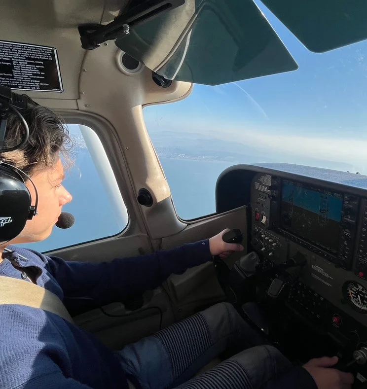 Pilot flying a small aircraft over the ocean with a view of the coastline under clear blue skies.