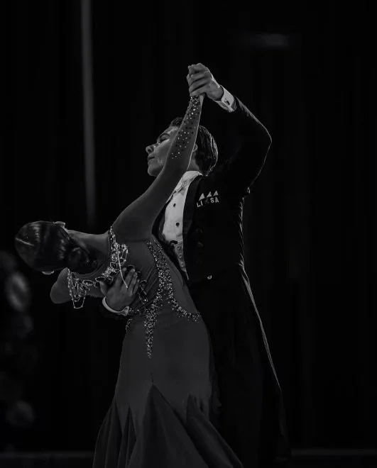Black and white photo of a couple performing a ballroom dance, with the woman wearing a decorated dress and the man in a tuxedo, dancing closely with their arms raised.