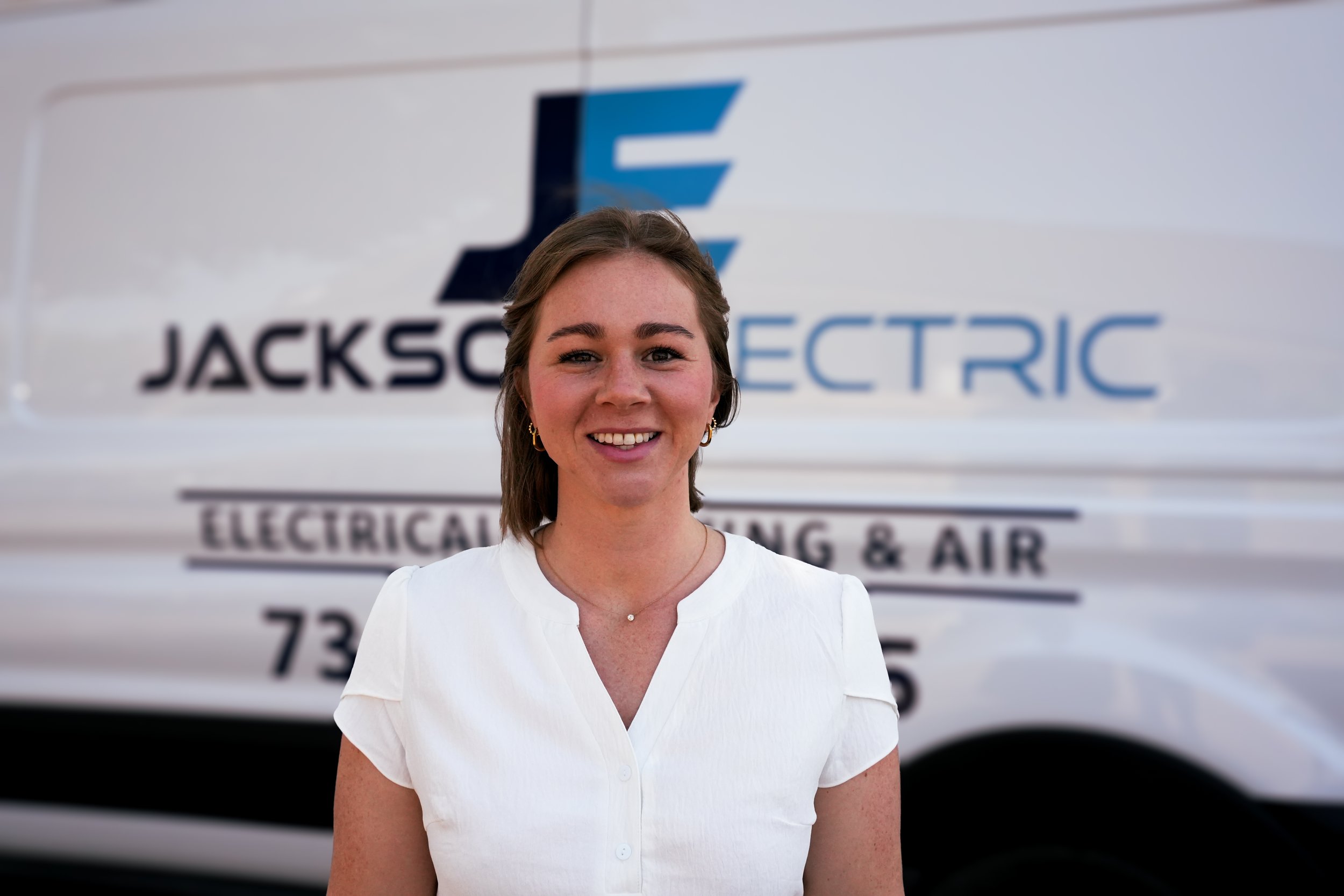 Smiling person with straight dark hair wearing a blue top in a leafy outdoor setting.