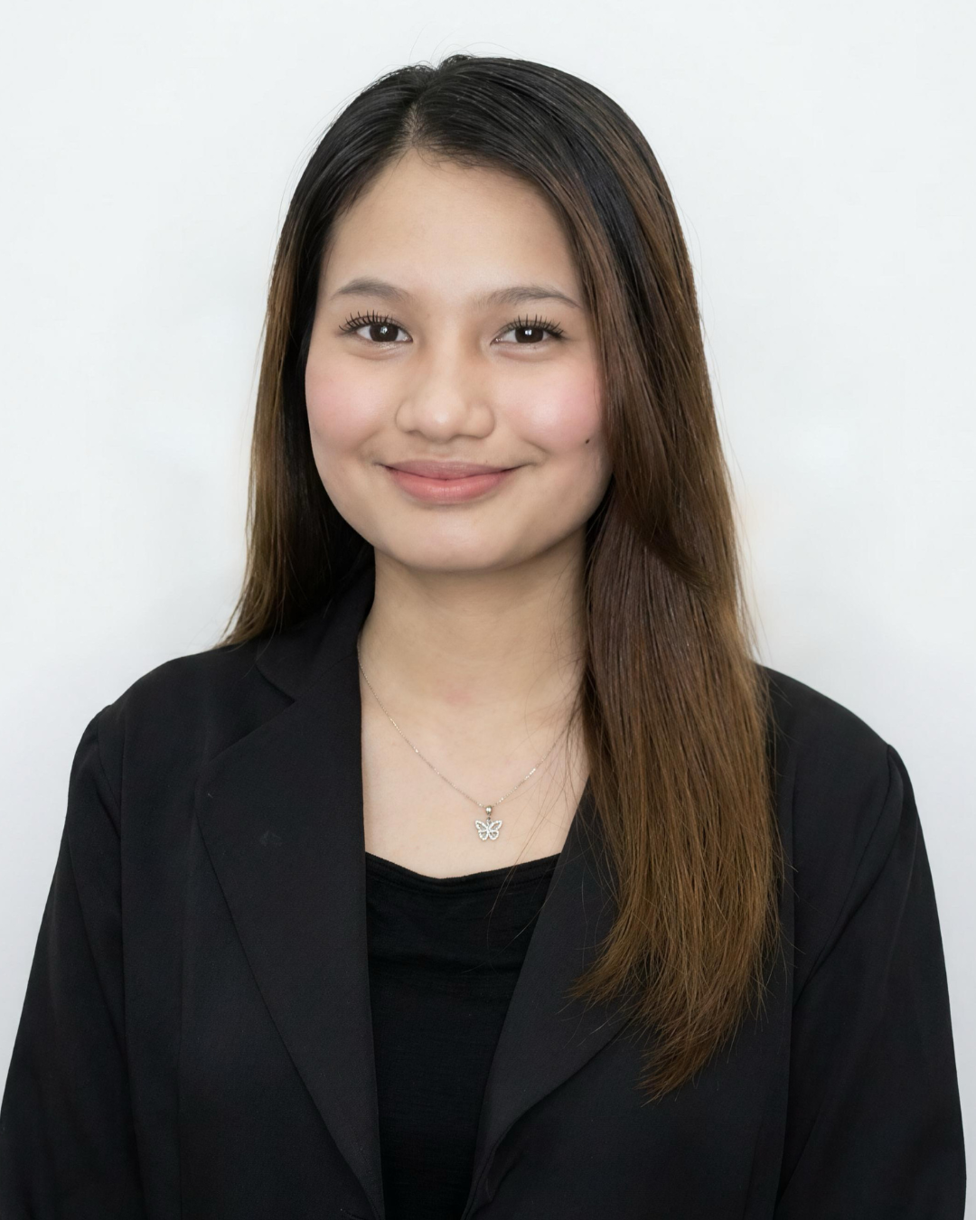 A smiling woman with long dark brown hair wearing a black blazer and black top, standing in front of a light-colored background.