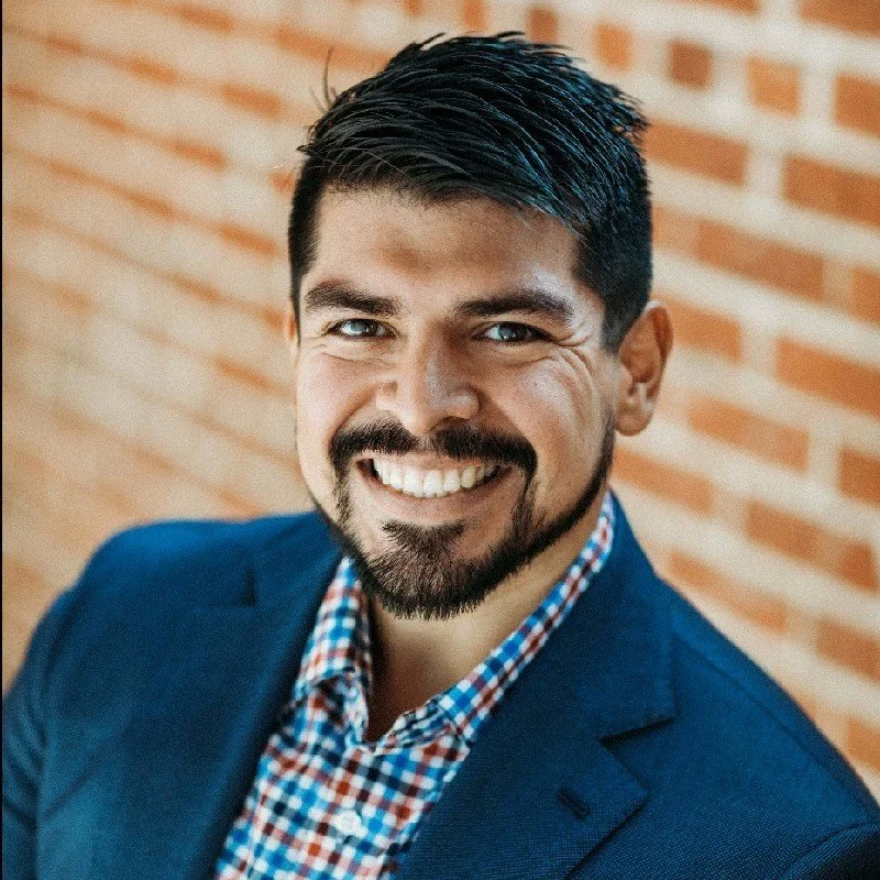 A reliable professional electrical engineer man with dark hair and a beard smiling, wearing a blue blazer and a checkered shirt, standing in front of a brick wall.