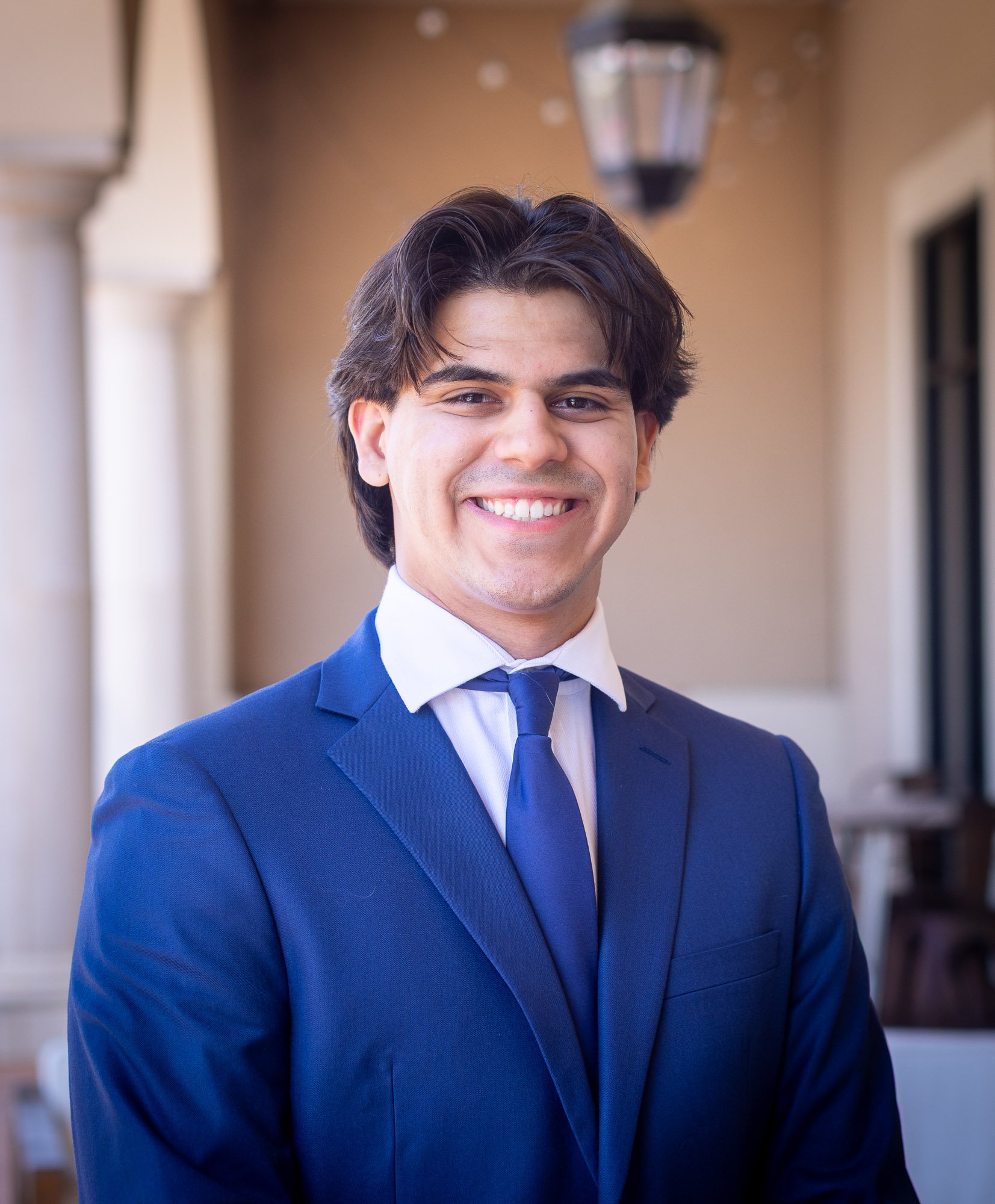 A smiling man with medium-length dark hair wearing a blue suit, white dress shirt, and blue tie, standing outdoors in front of a building.