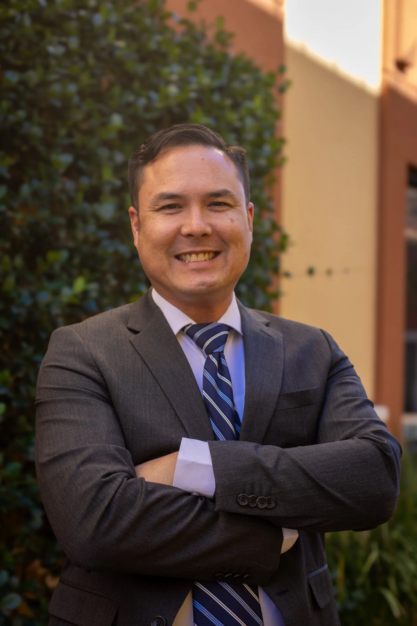 A smiling man with short dark hair wearing a charcoal gray suit, a light blue dress shirt, and a blue striped tie, standing outdoors with his arms crossed in front of a building and greenery.