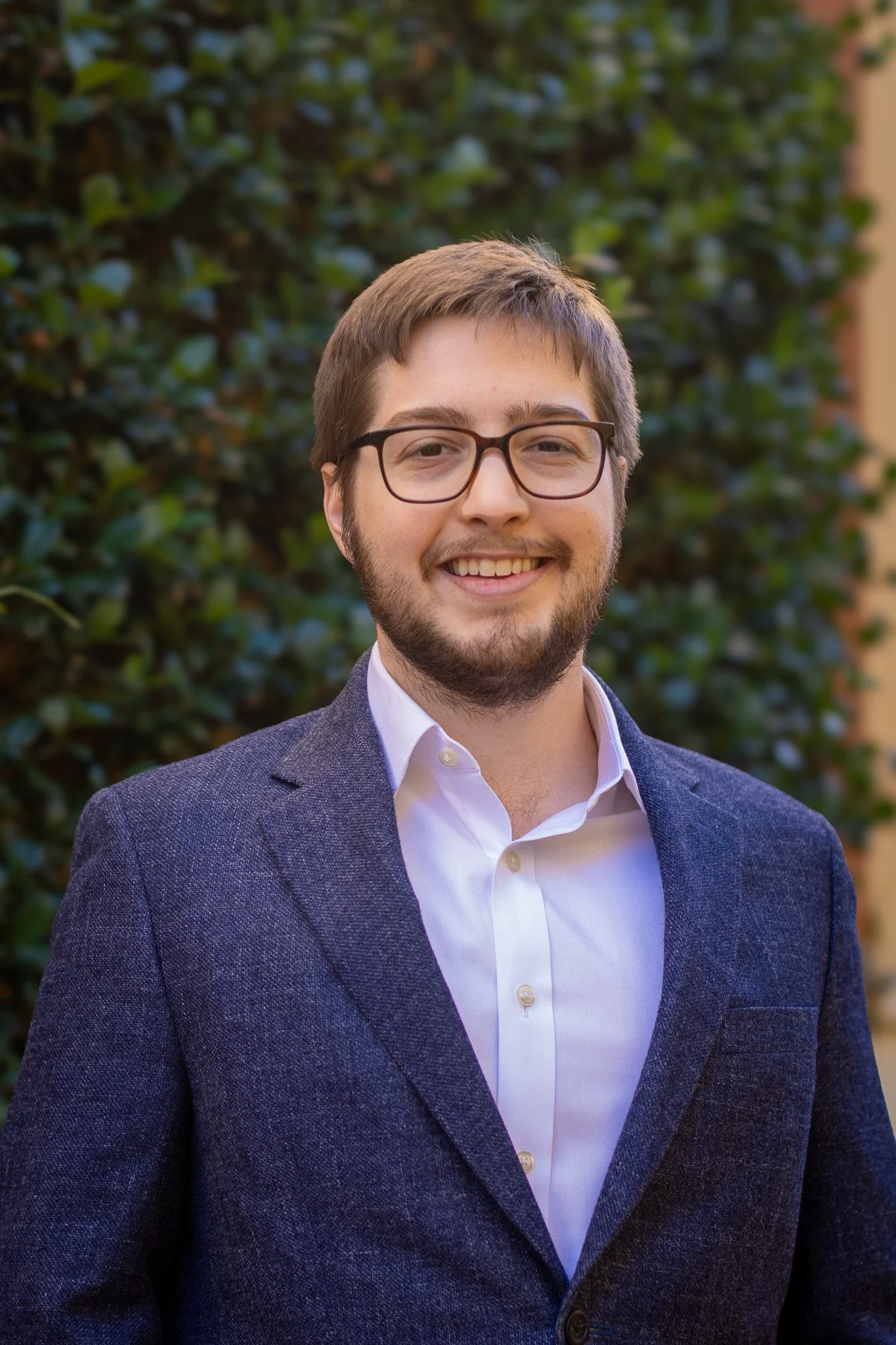 A smiling man with short brown hair, glasses, and a beard wearing a textured navy blazer and a white dress shirt, standing outdoors in front of green trees.