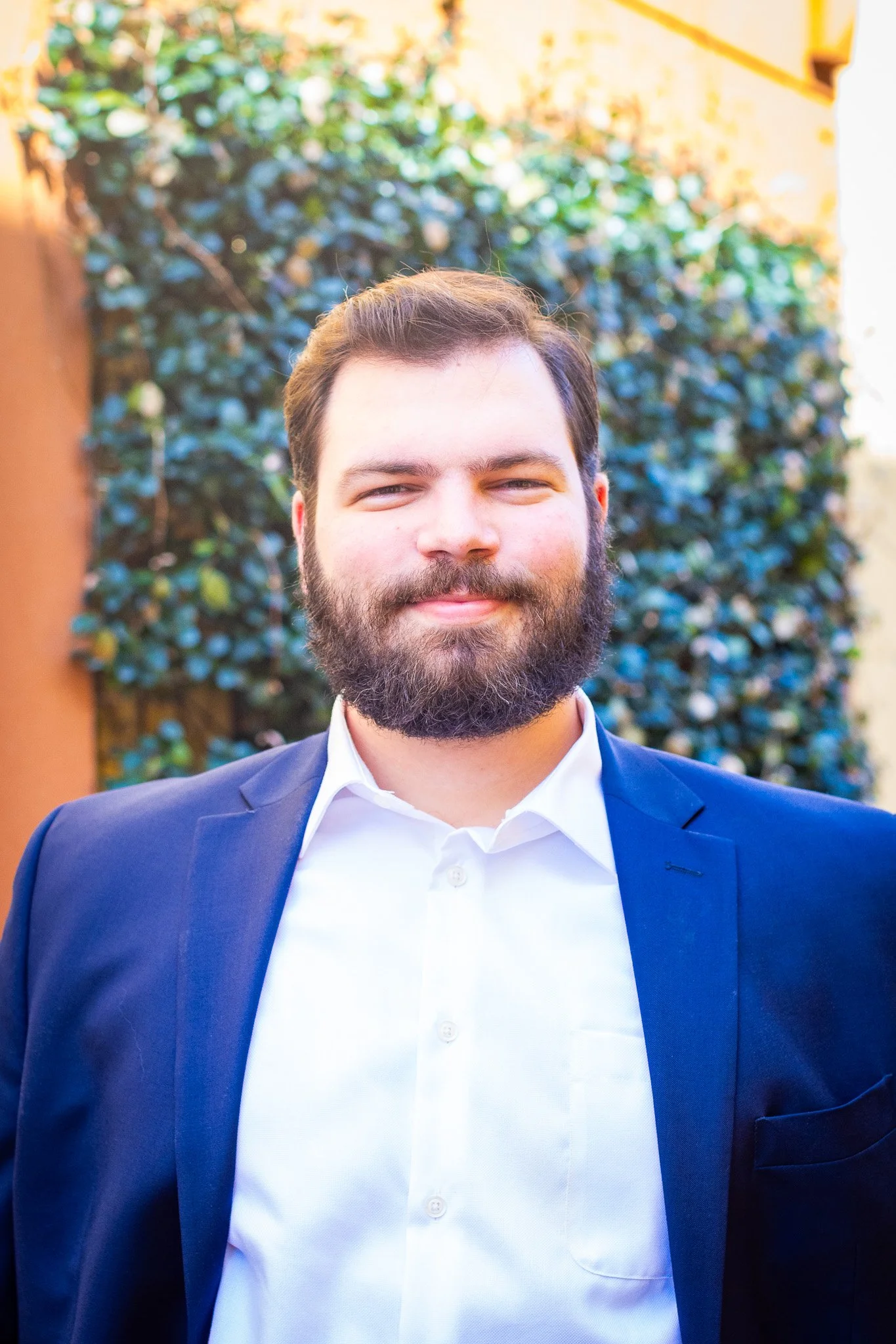 A smiling man with dark hair and a full beard wearing a navy blazer and a white dress shirt, standing outdoors in front of greenery and a building.