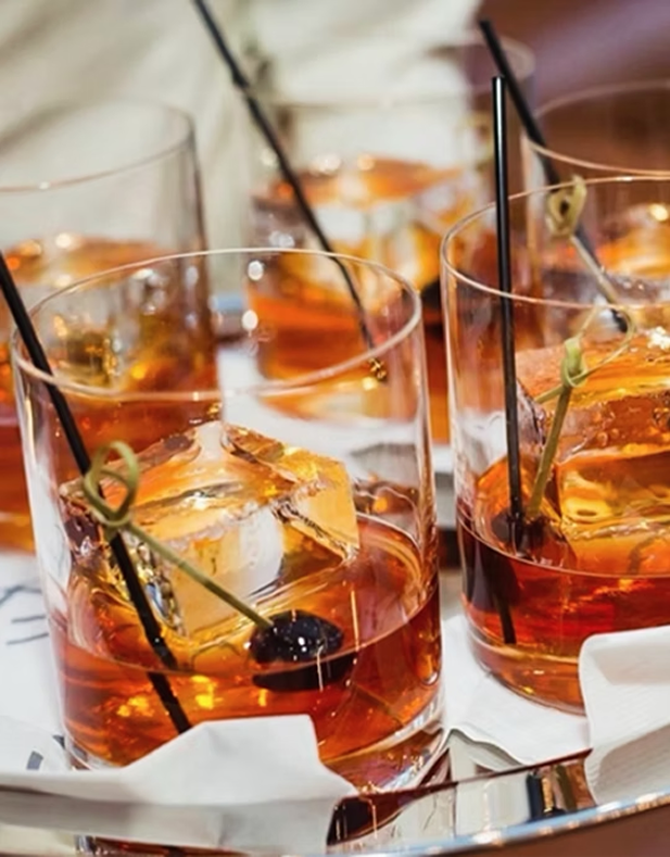 Several glasses of bourbon with club soda, ice, and a cherry garnis, served on a silver tray with a white cloth.