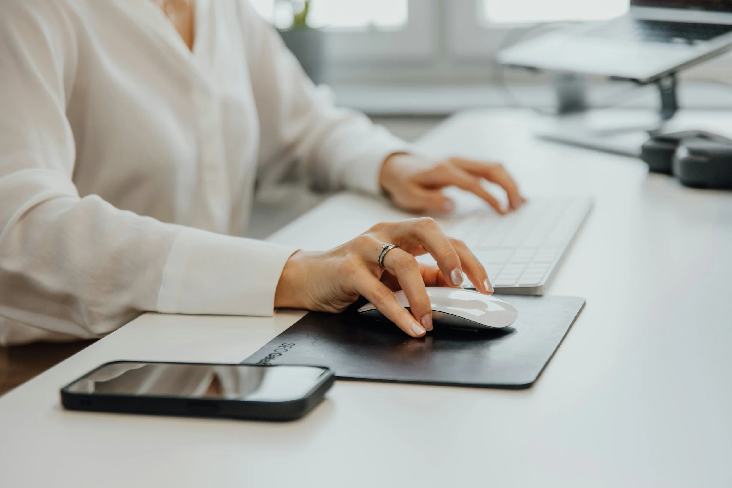 Person in a white blouse working on a computer at a desk, with a smartphone nearby.