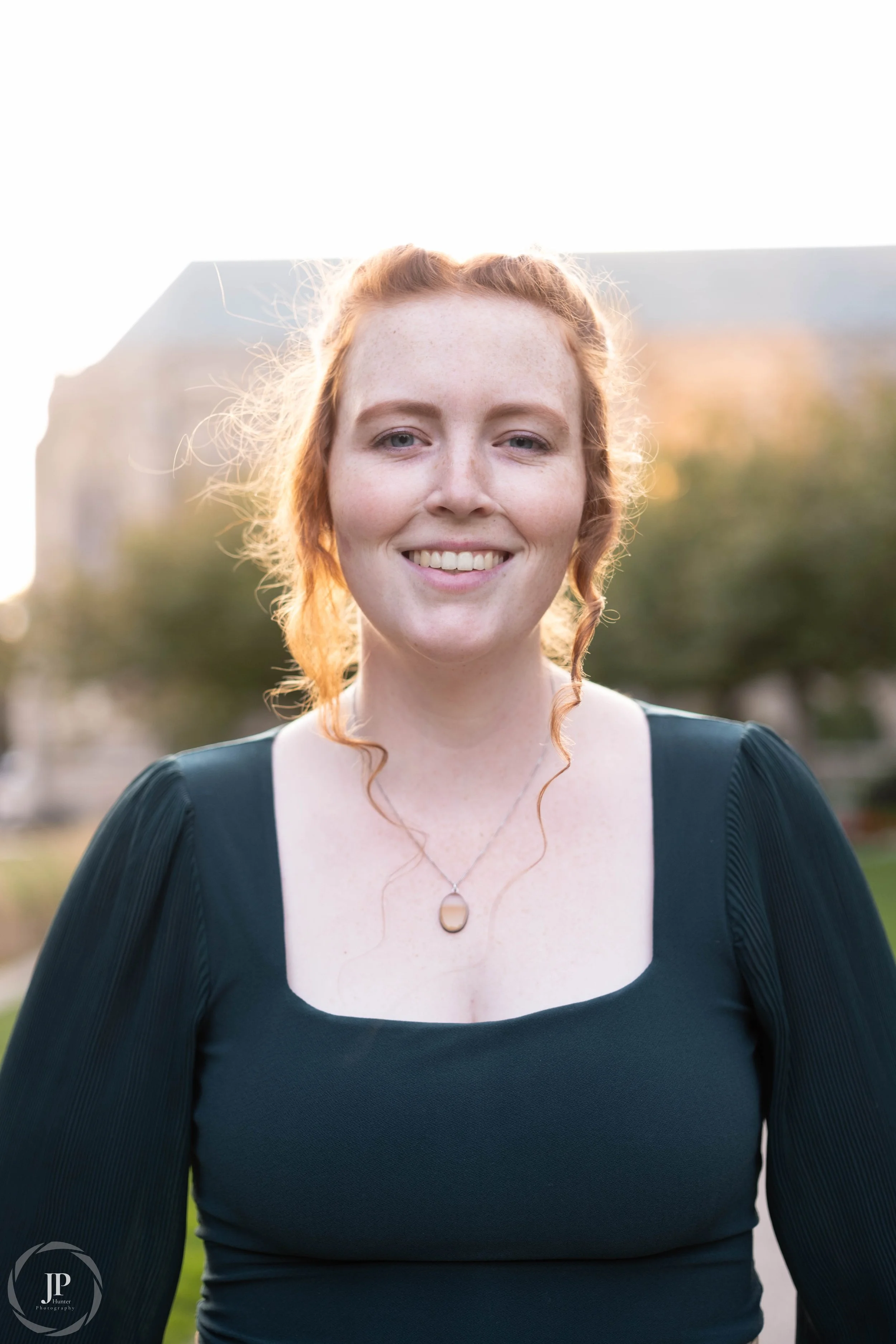 Woman smiling outdoors, wearing a green dress with necklace, backlit by sunset.