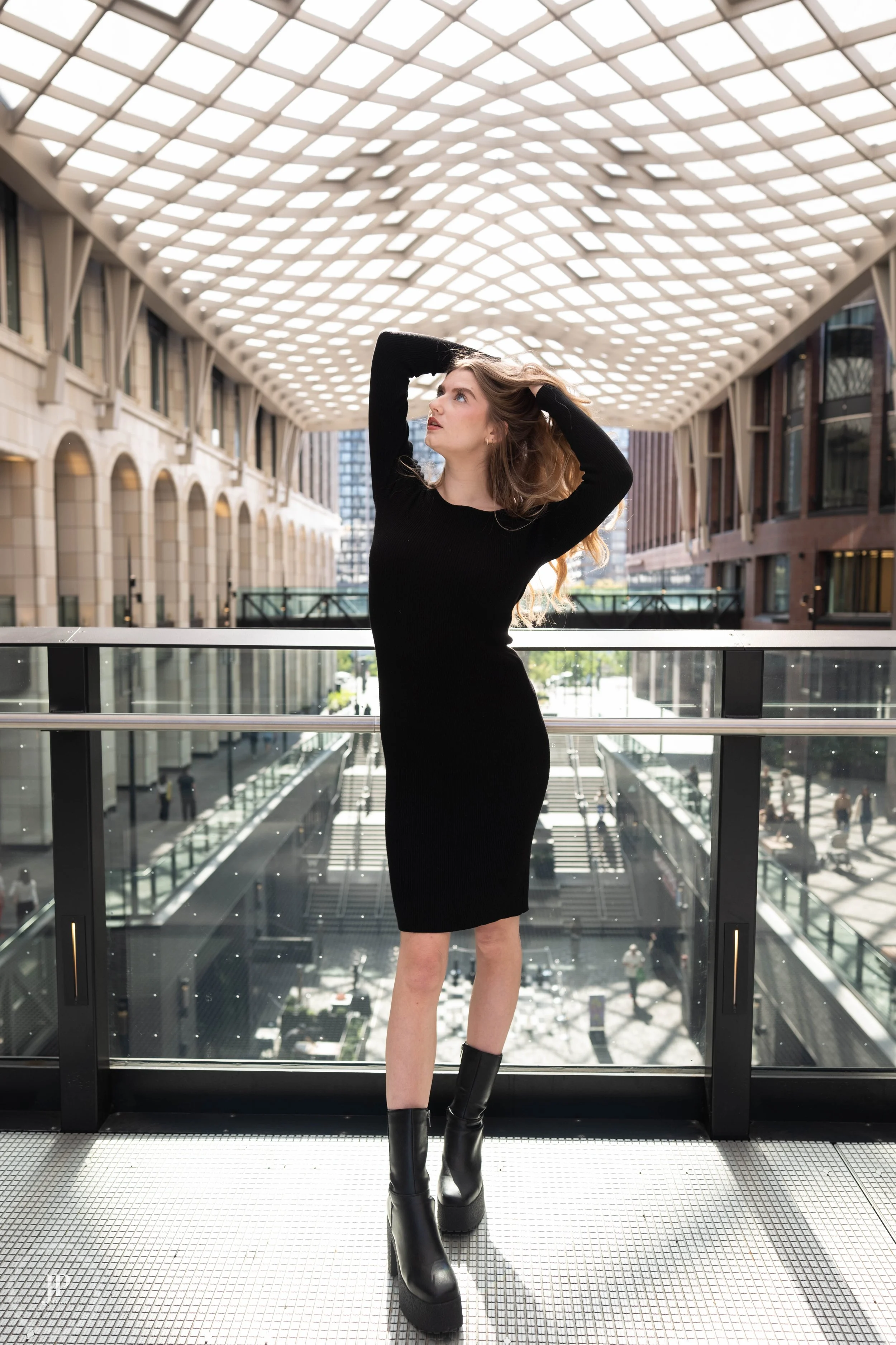 Woman in black dress and boots posing in a modern atrium with a geometric ceiling.