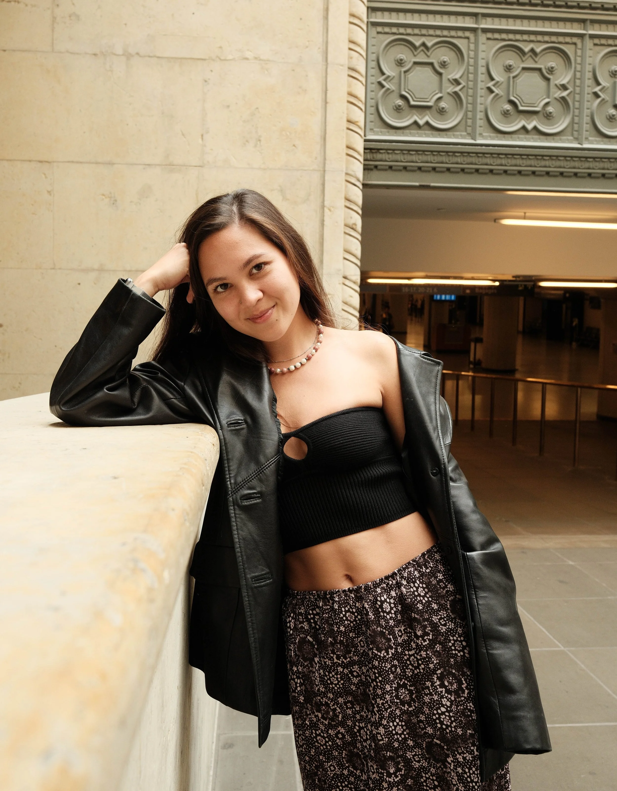 Woman posing in a stylish black outfit, leaning on a ledge in a decorative indoor setting.