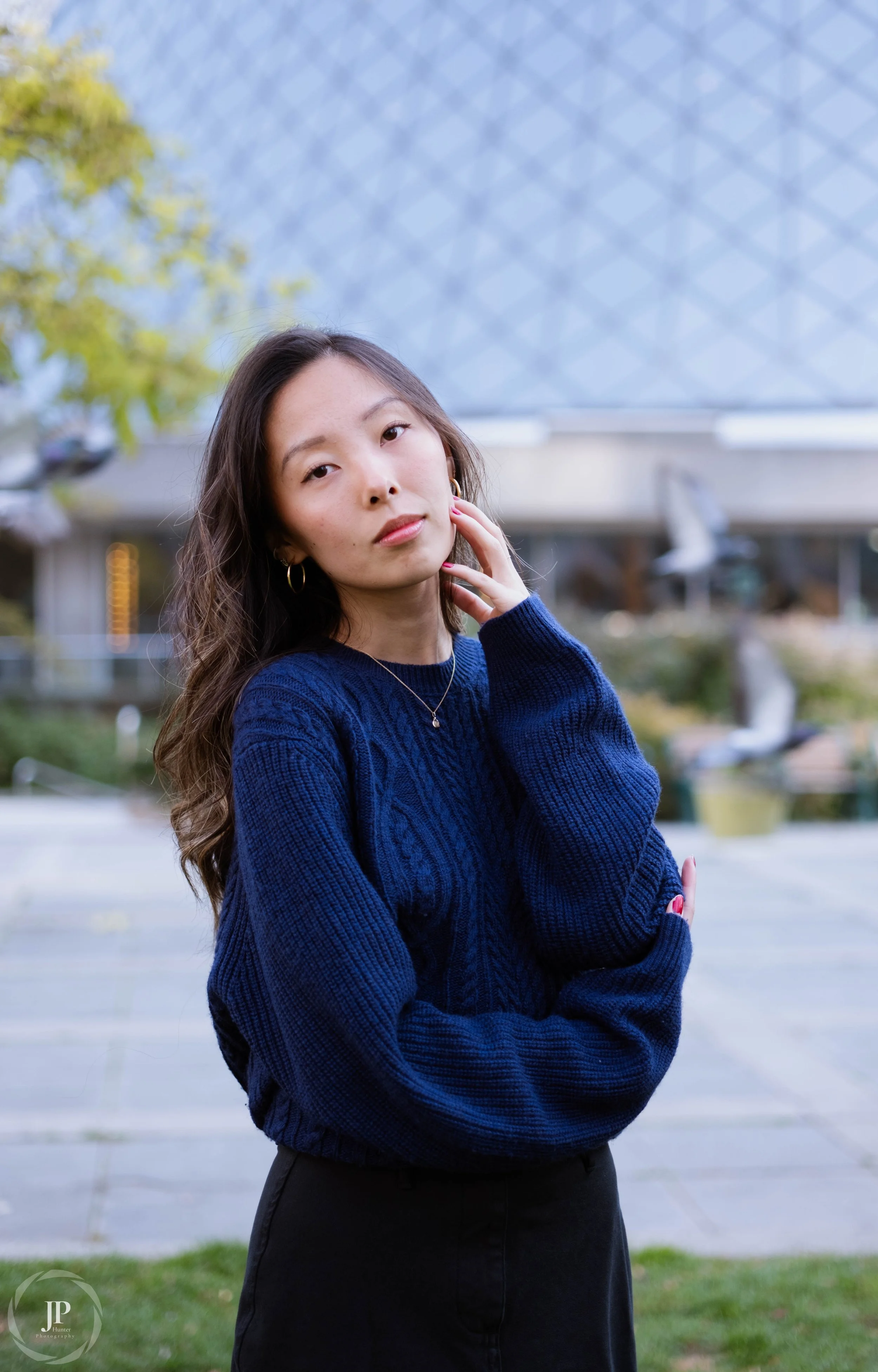 Woman wearing a blue sweater, standing in an outdoor setting with a building in the background, touching her face gently.