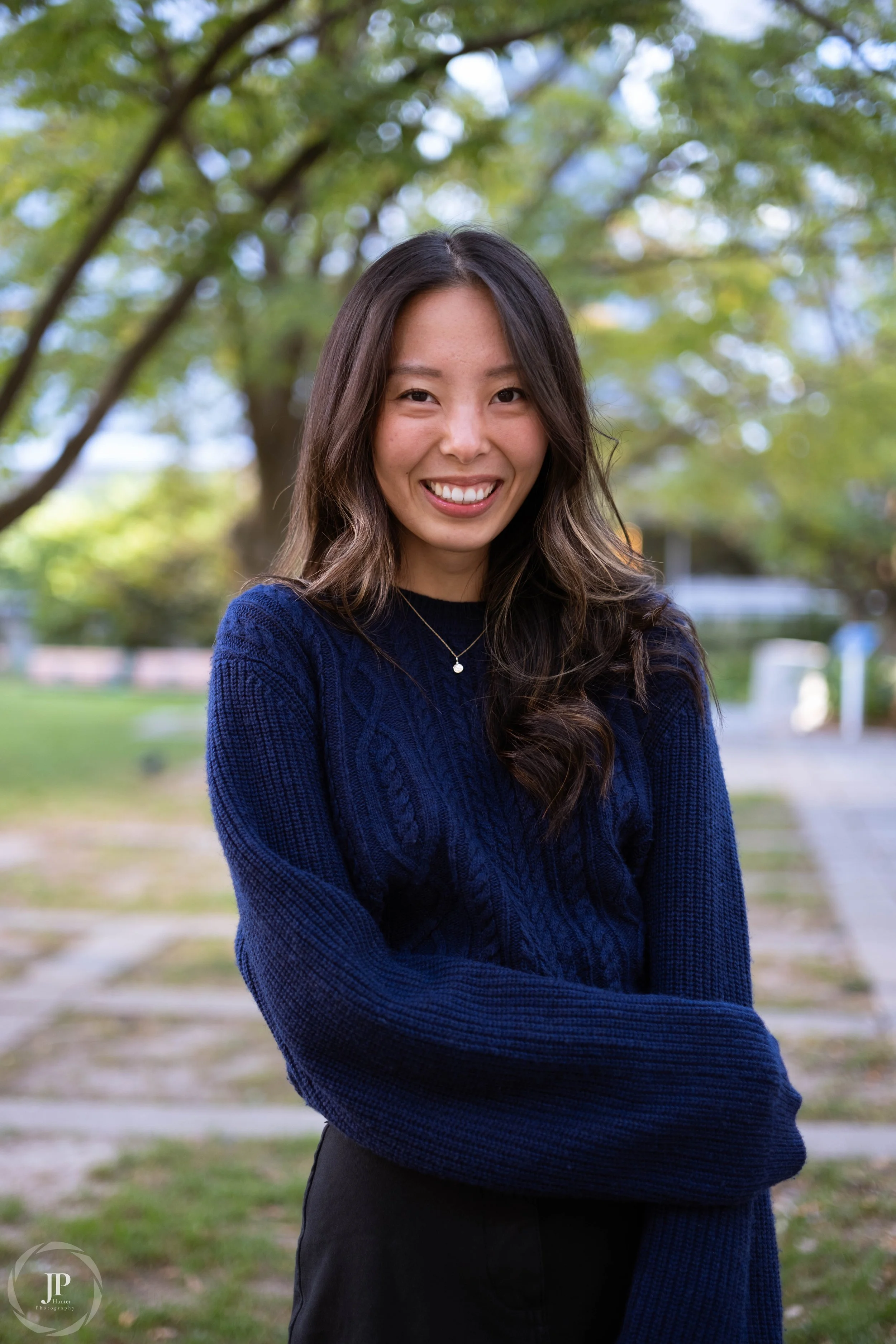 Woman in a blue sweater standing outdoors with trees in the background, smiling.