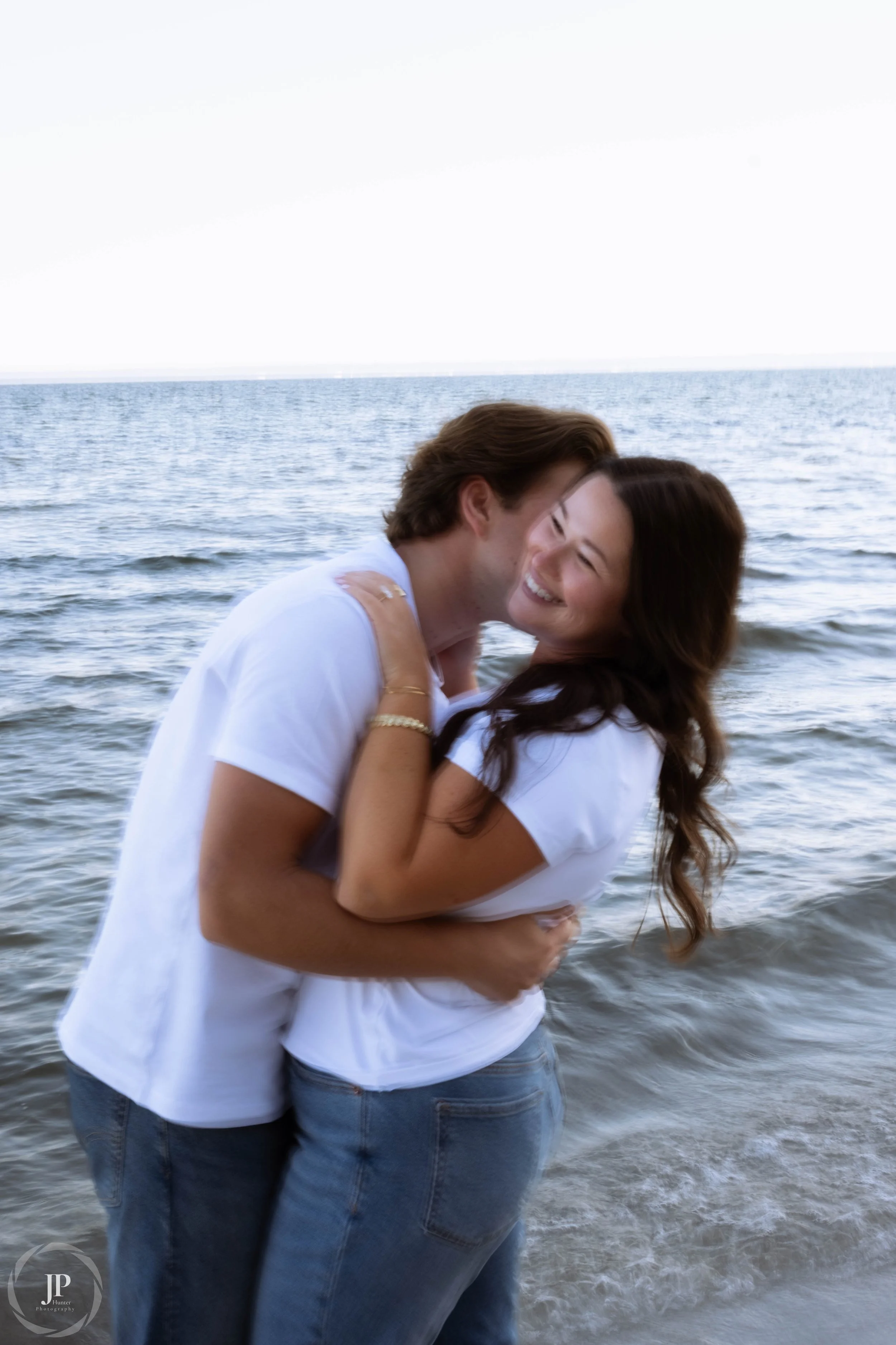 Couple embracing by the ocean, both wearing white shirts and jeans, with the sea in the background.