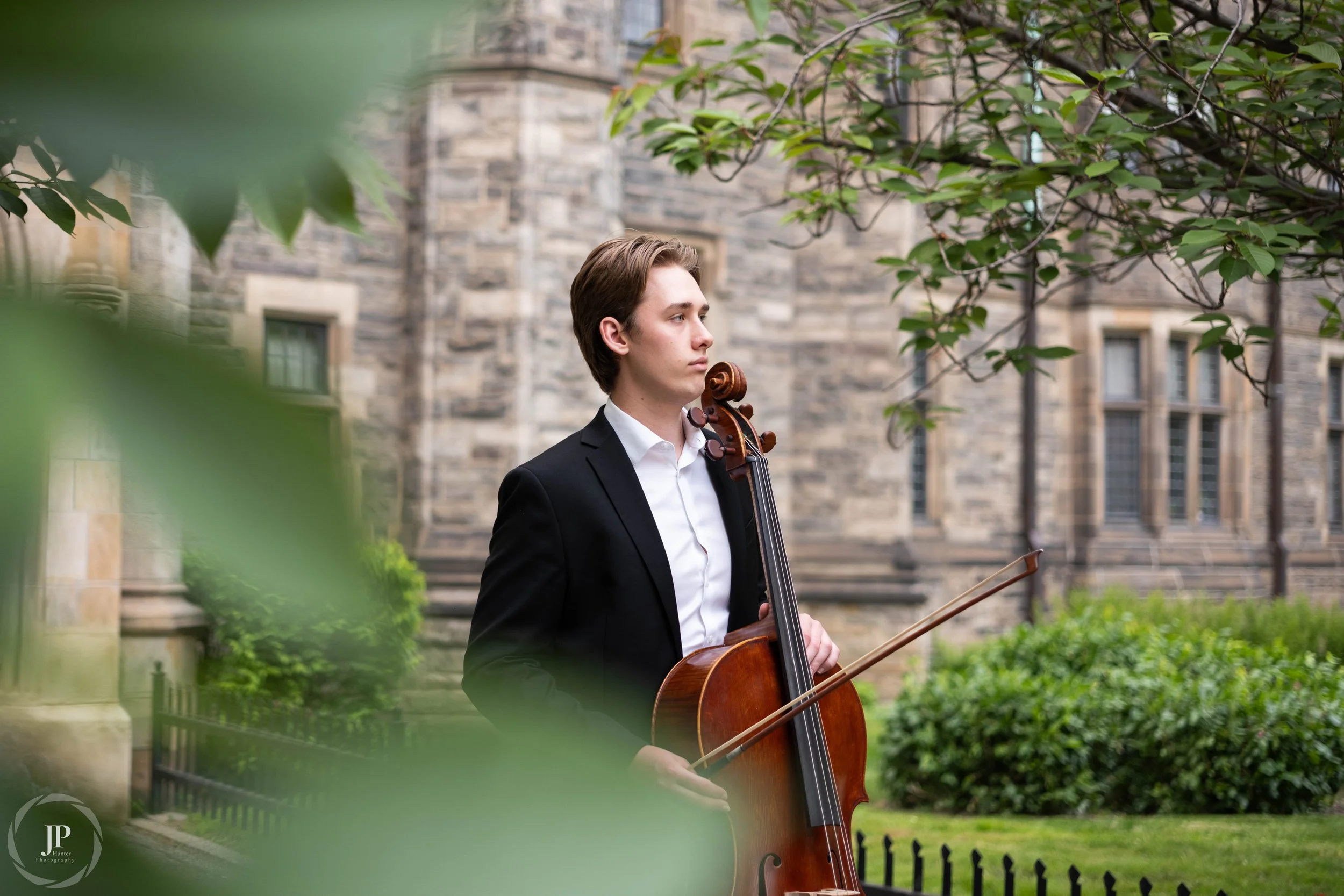 A person posing with his Cello in a suit with artistic leaves in the foreground and stonework in the background