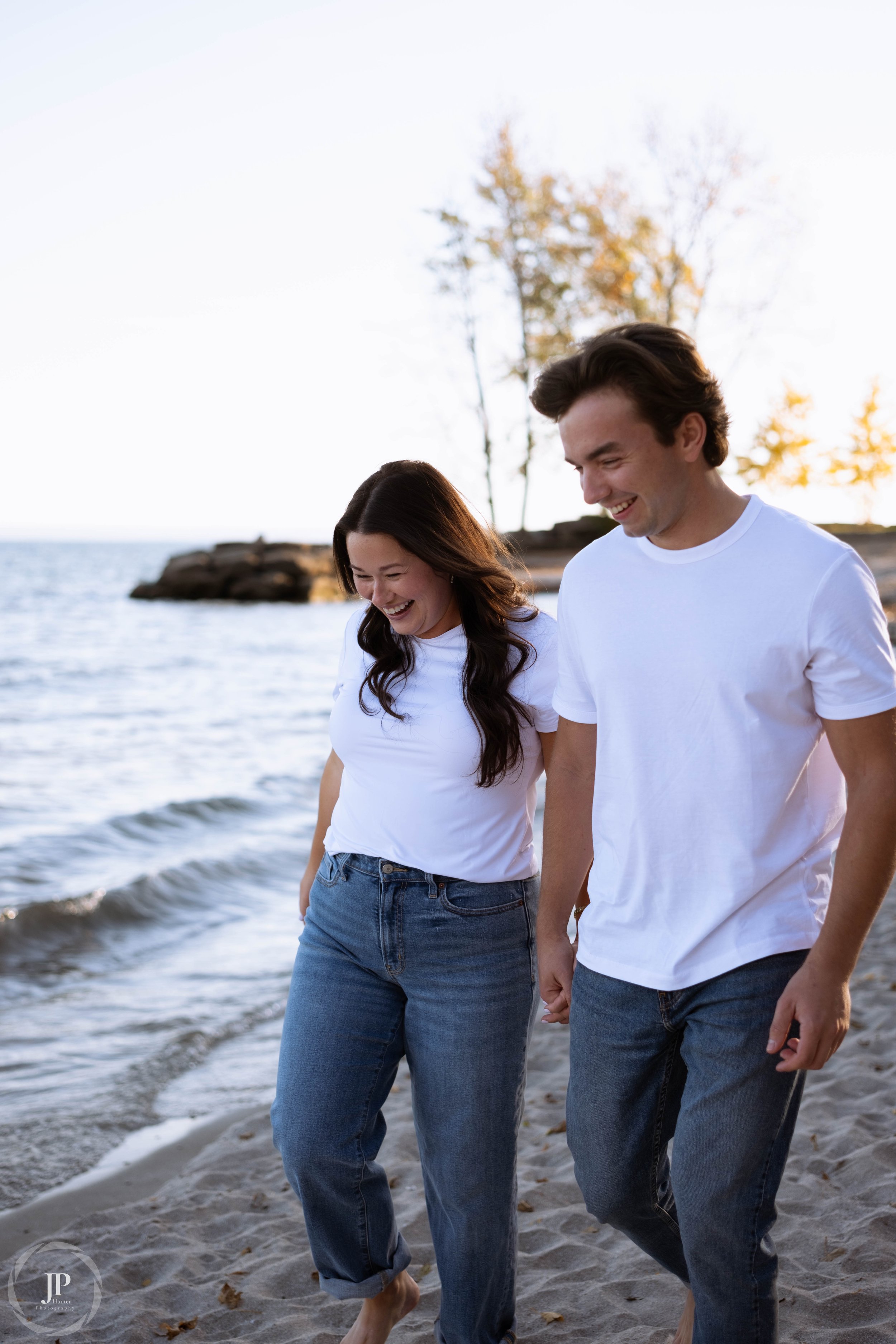 A smiling couple in white t-shirts and jeans walking on a beach near the water.