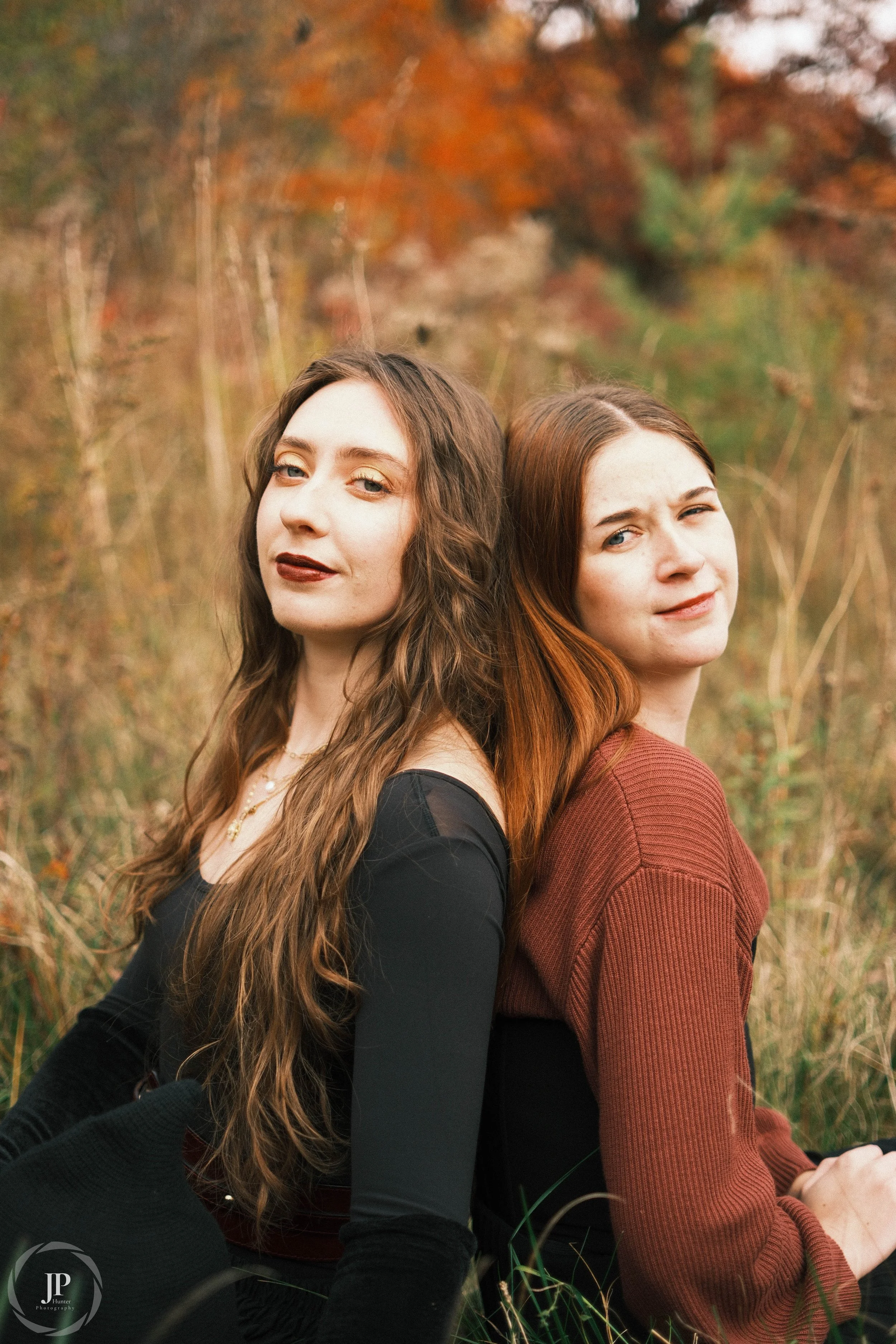 Two women sitting back-to-back in a grassy field during autumn