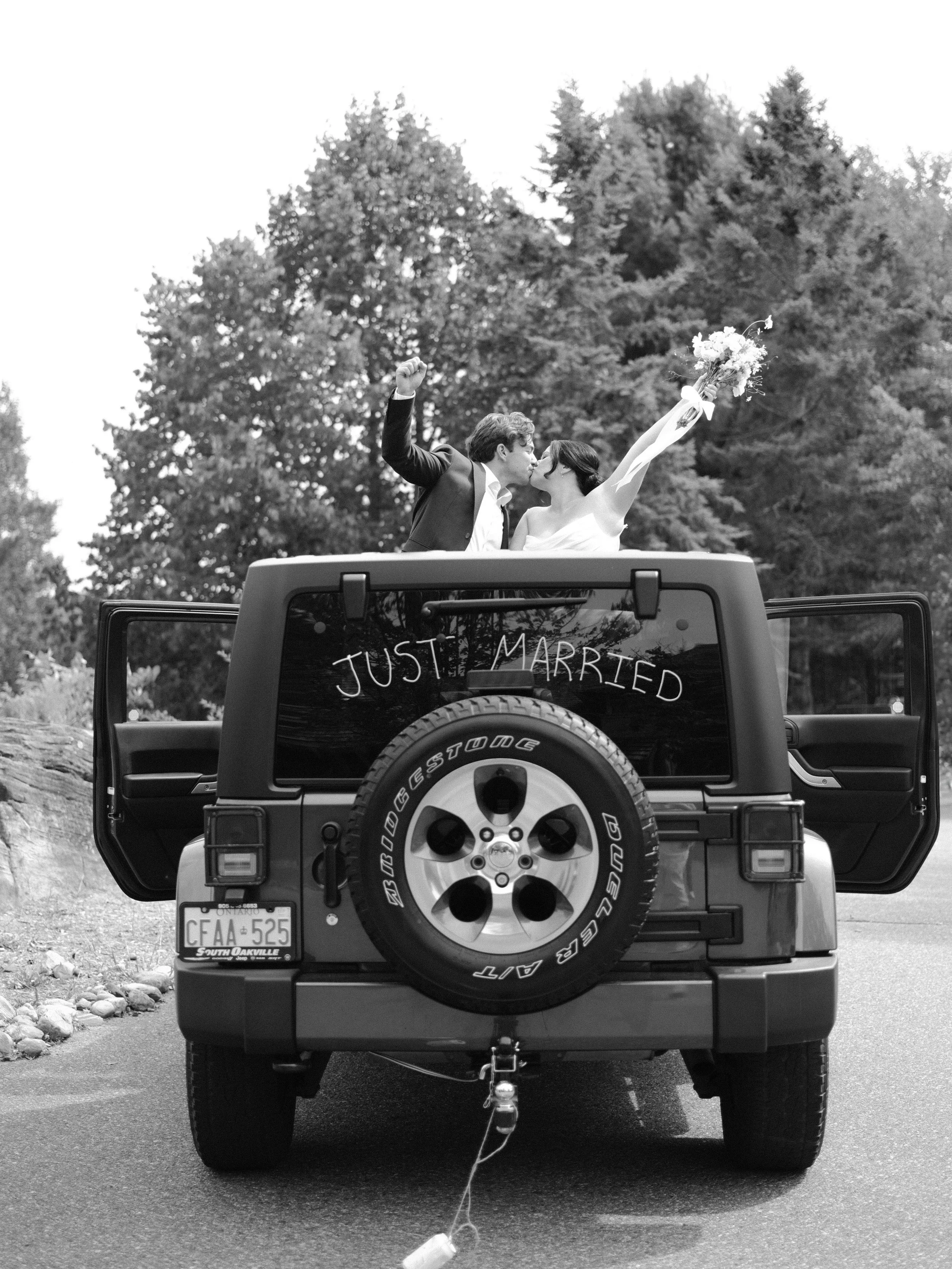 A just married couple cheering out of a jeep that has "just married" written on the car with cans trailing behind