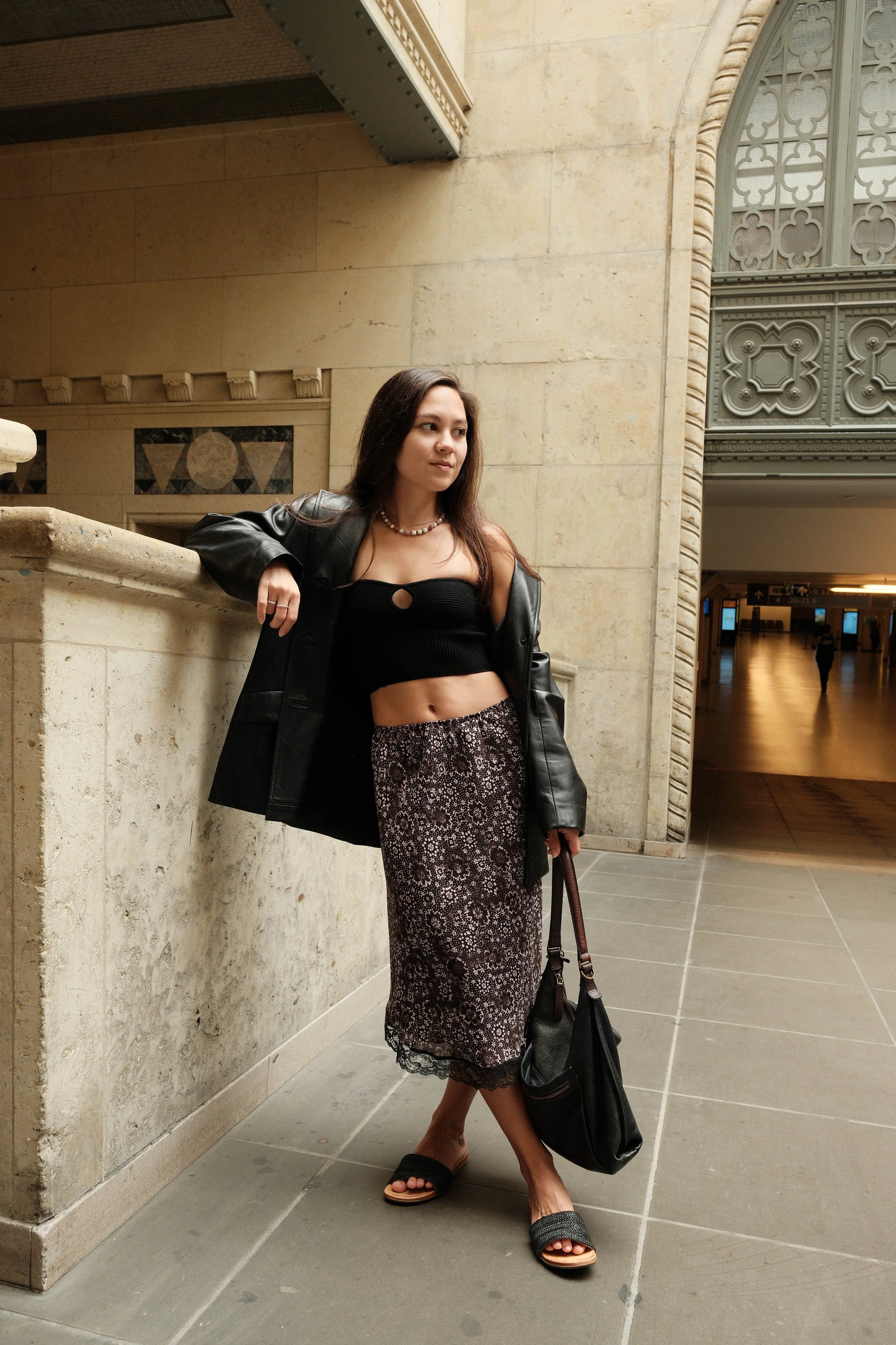 Woman in black crop top and patterned skirt leaning on stone railing indoors.