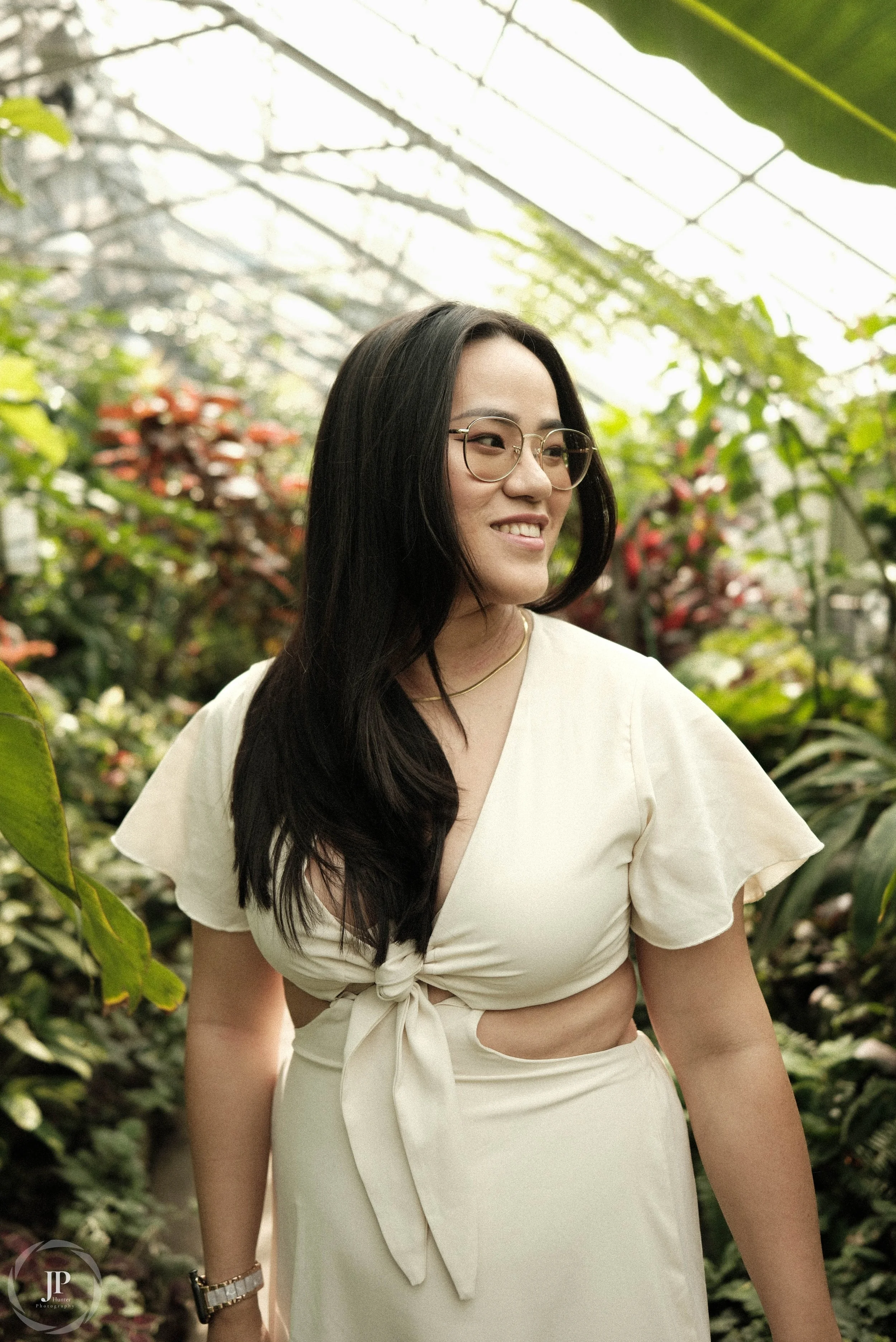 Woman in a light-colored dress standing in a greenhouse surrounded by lush plants.