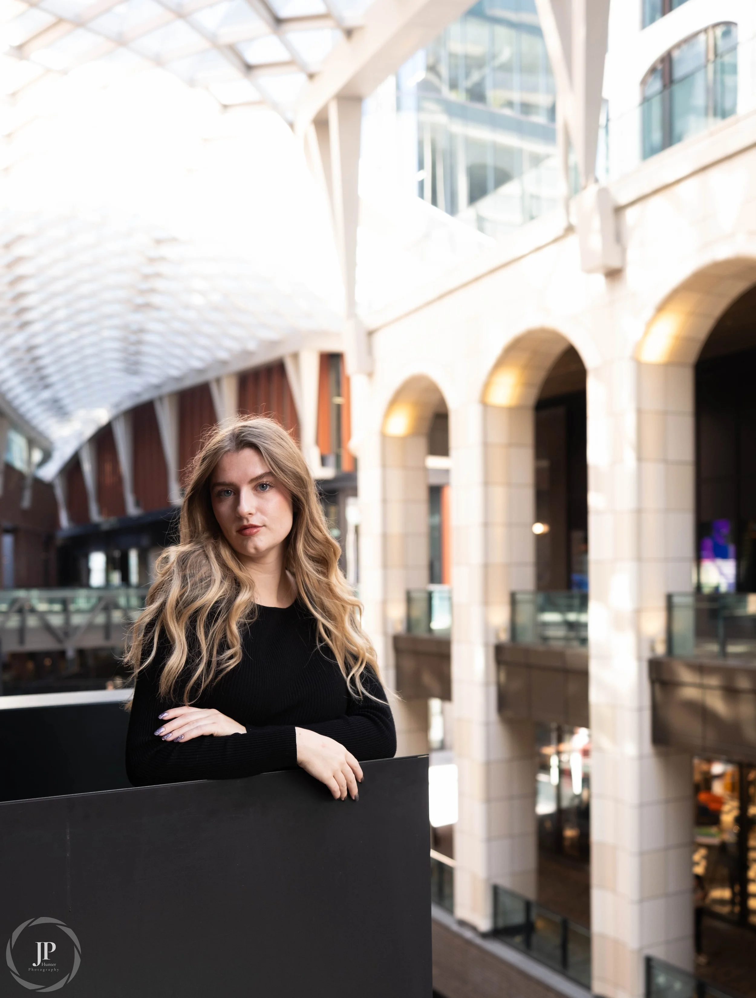 Woman in black outfit posing indoors with modern architecture in background.