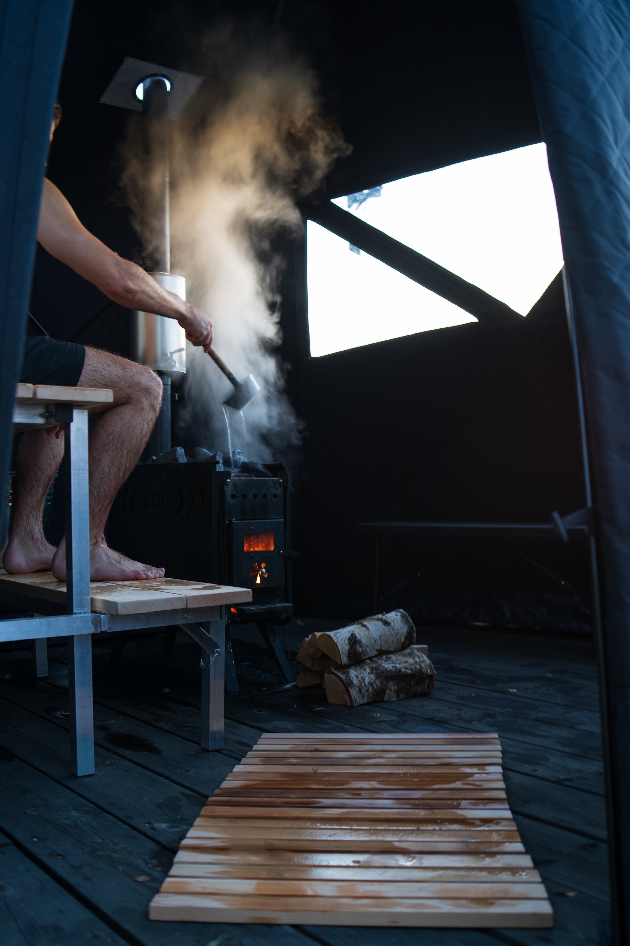 Person inside a sauna pouring water on a wood-burning stove, with steam rising. The floor is wooden with scattered logs next to the stove.