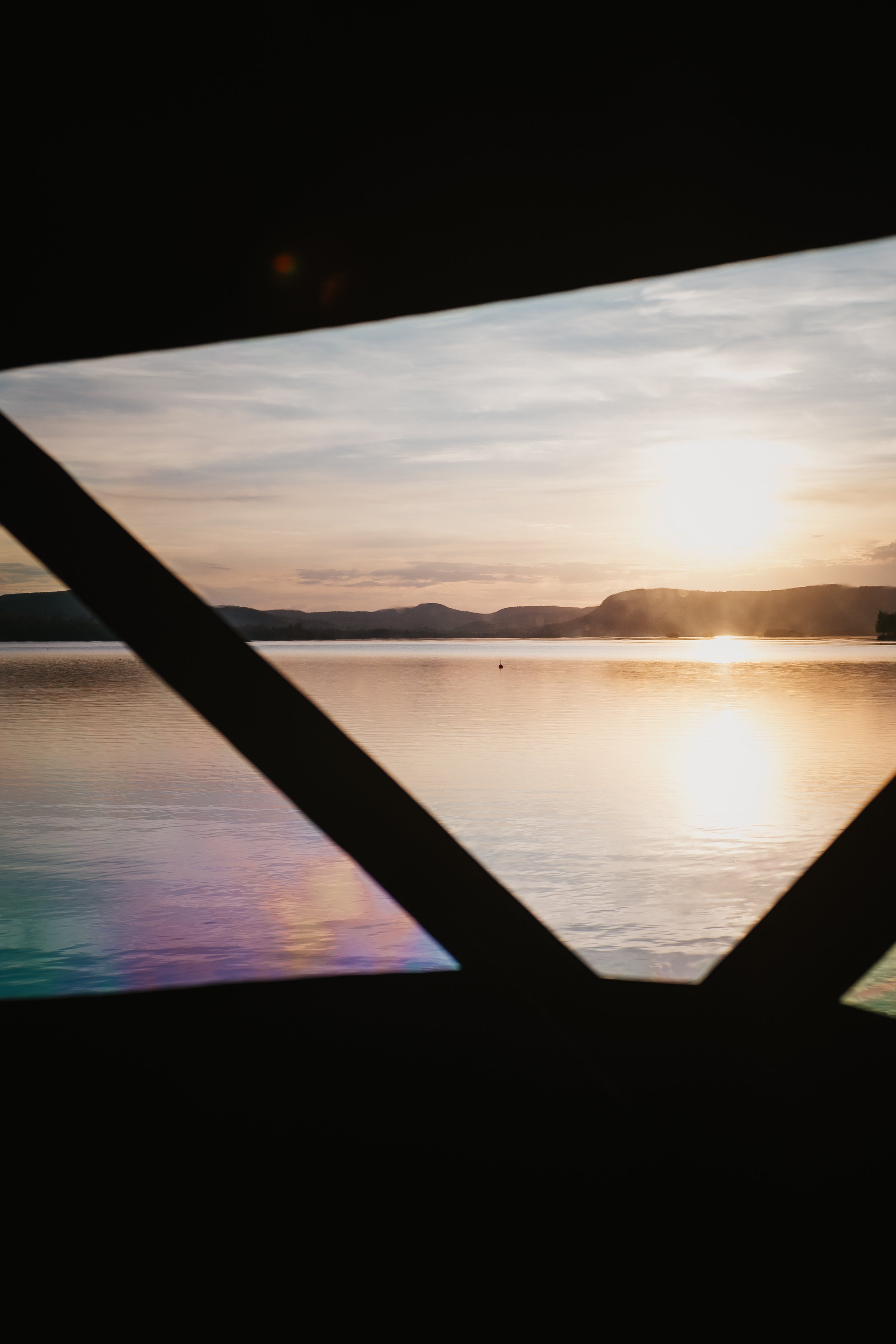 Sunset over a calm lake viewed through a window with crossbars, reflecting warm colors on the water's surface.
