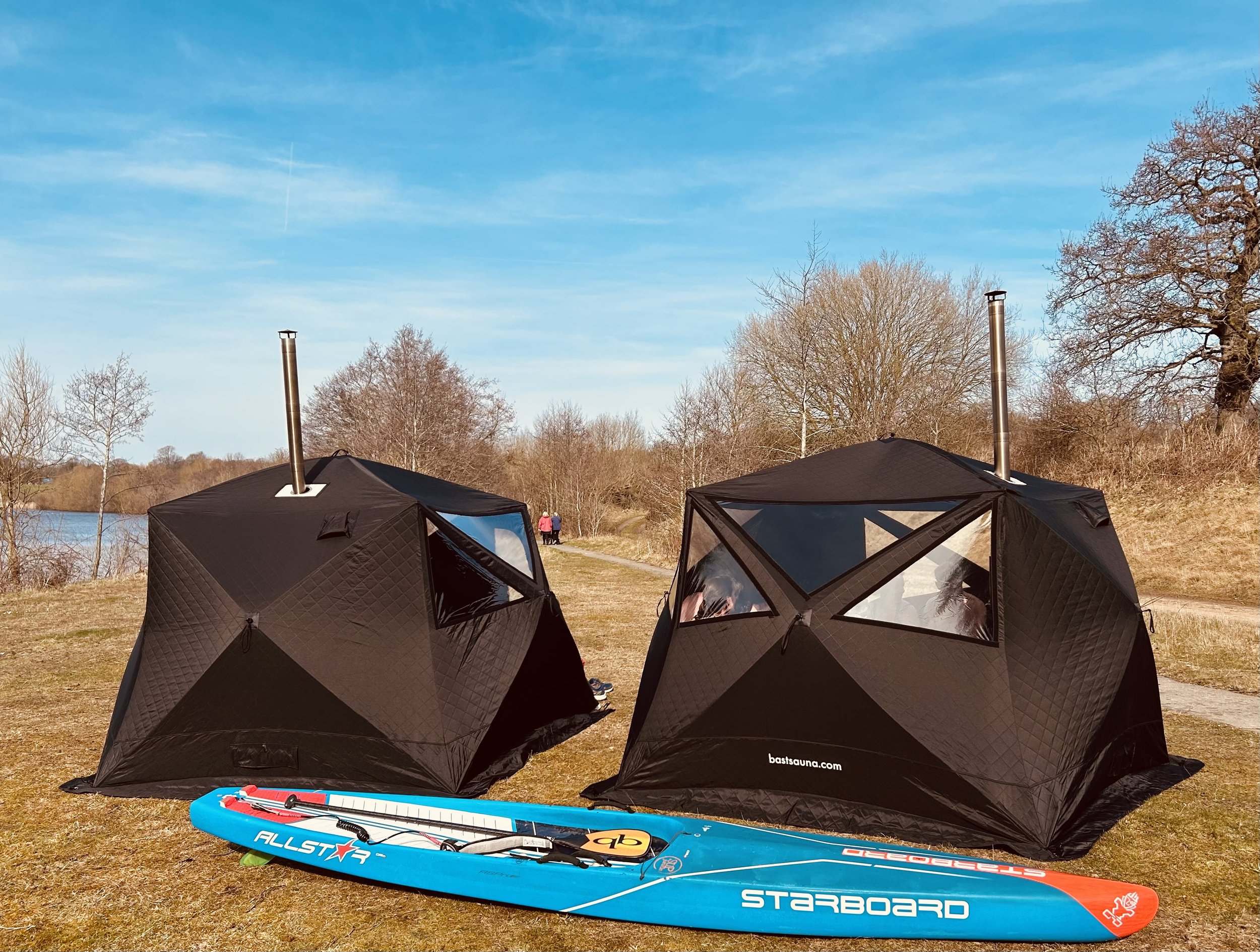 Two black saunas with chimneys set up outdoors on grassy terrain with a blue and red paddleboard in front, next to a path and trees under a blue sky.