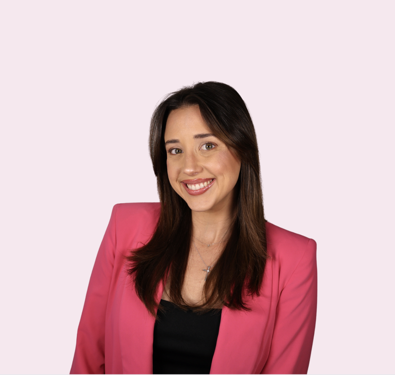 A woman with long dark hair smiling with crossed arms wearing a pink blazer over a black top against a light pink background.
