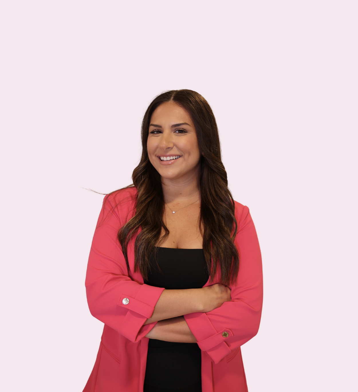 A woman with long brown hair, wearing a black top and a pink blazer, smiling and crossing her arms against a light pink background.