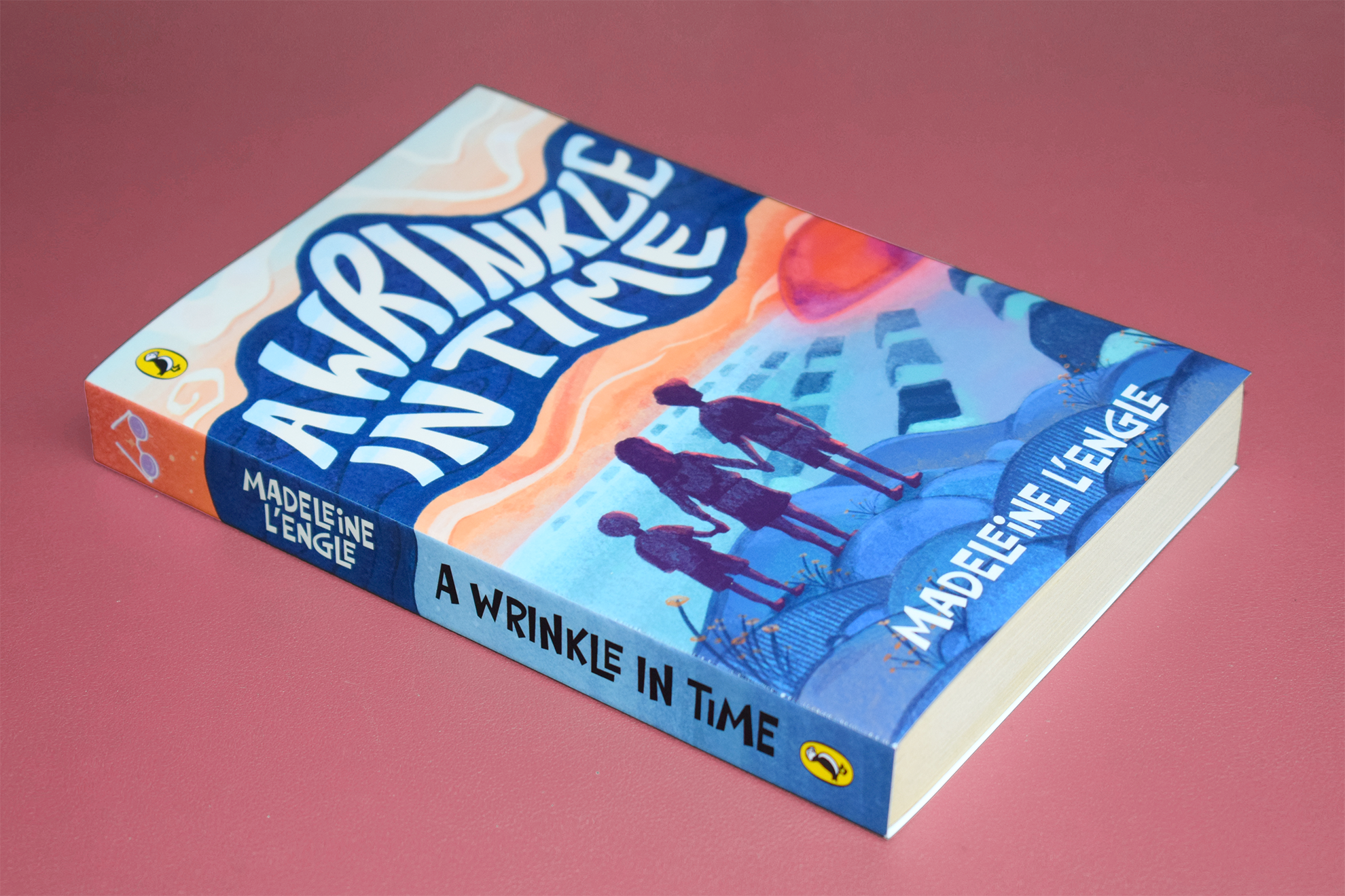 Book titled 'A Wrinkle in Time' by Madeleine L'Engle lying on a pink surface. The cover features an illustration of three children with a landscape of abstract shapes and colors.