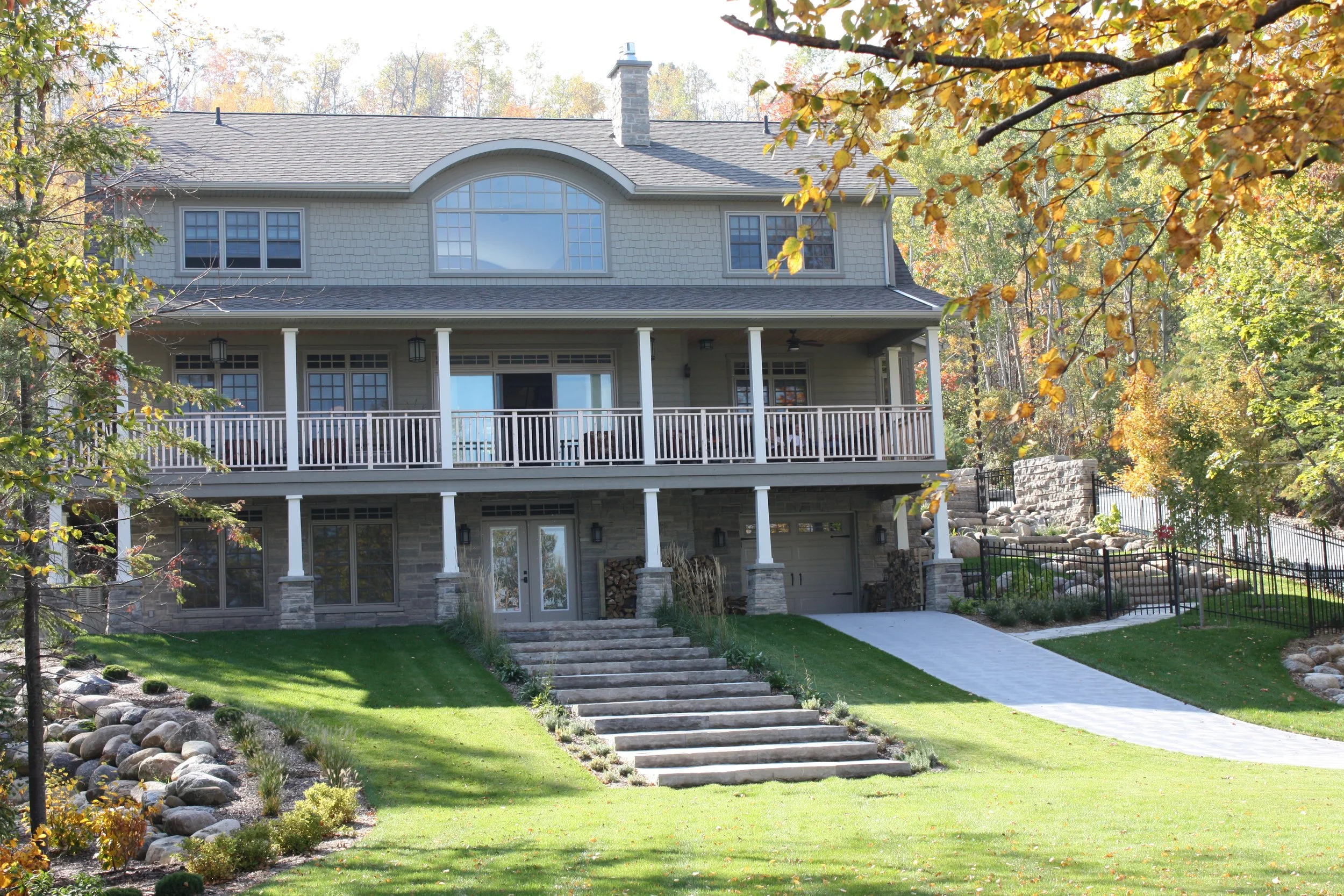 A large two-story house with a porch and balcony, surrounded by autumn trees and landscaping with stone steps leading to the entrance.